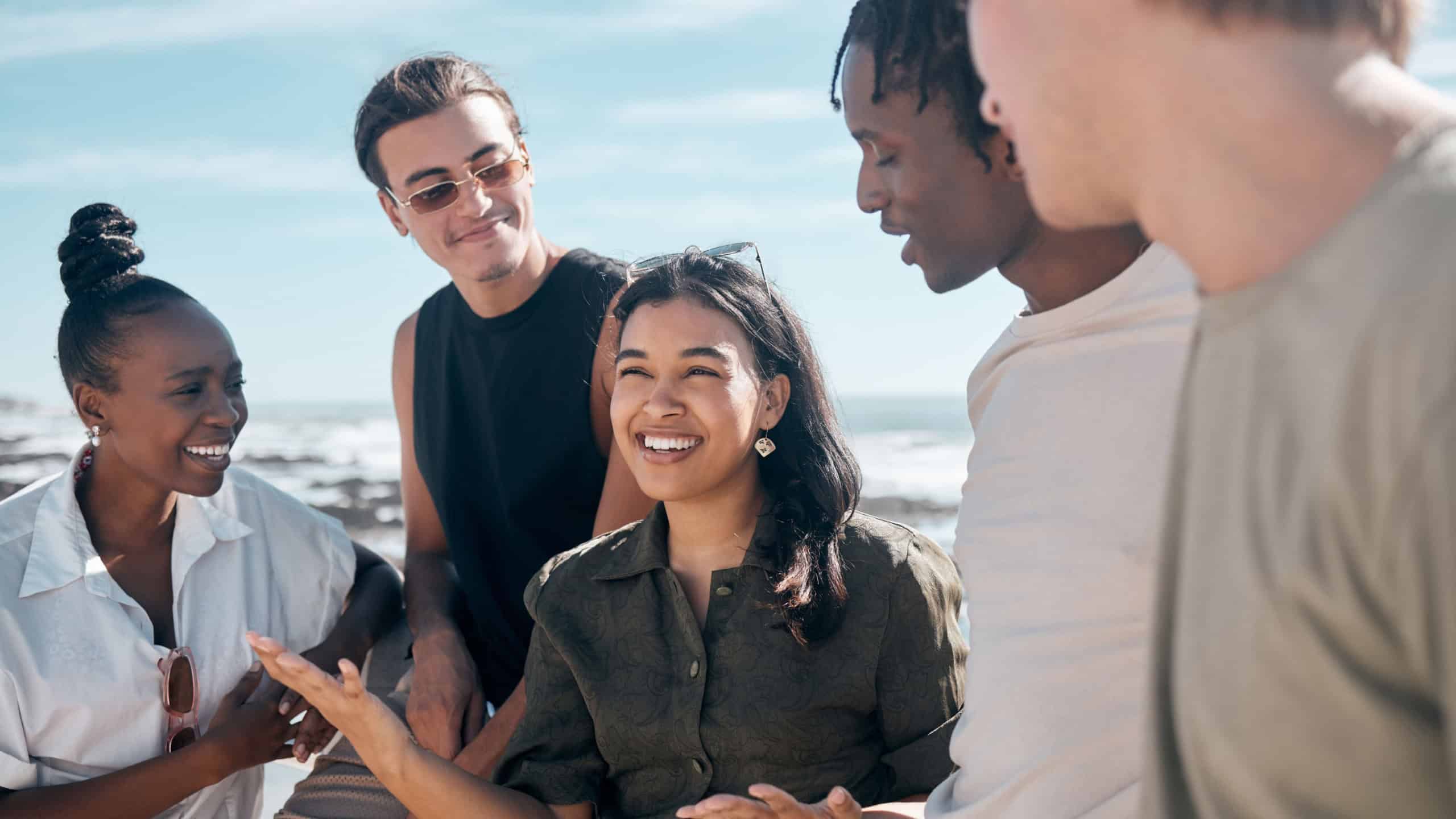 Woman, talking and bonding people by beach, ocean or sea in group social gathering, holiday community or summer storytelling. Smile, happy and diversity friends in travel location, relax or freedom
