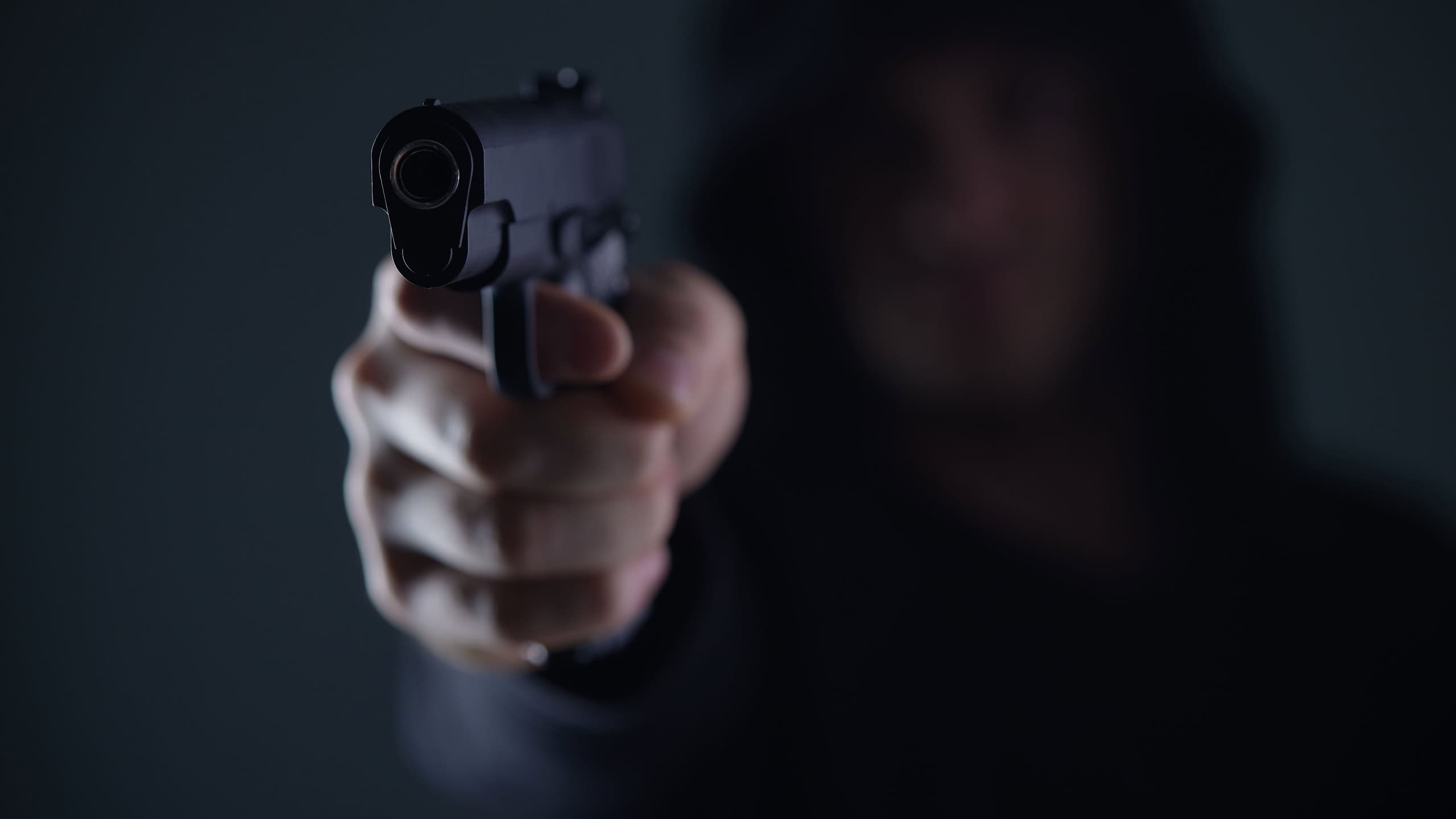 robber with gun aiming into camera. Man in hood threatens with firearm. Weapon in person's hands. Murderer or armed thief. Criminal with pistol.