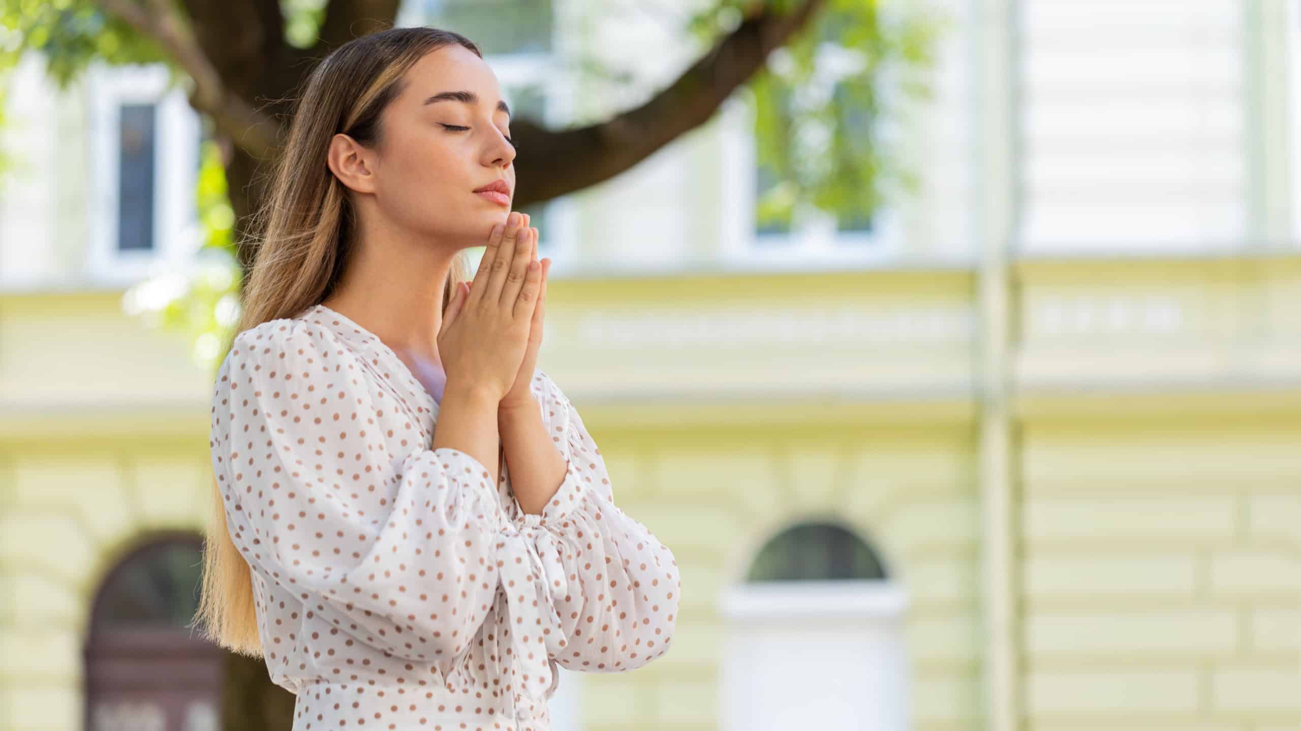 Portrait of young woman praying with closed eyes to god asking for blessing, help, forgiveness outdoors. Pretty girl clasping hands wishing luck in sunny urban city street. Town religion lifestyles.