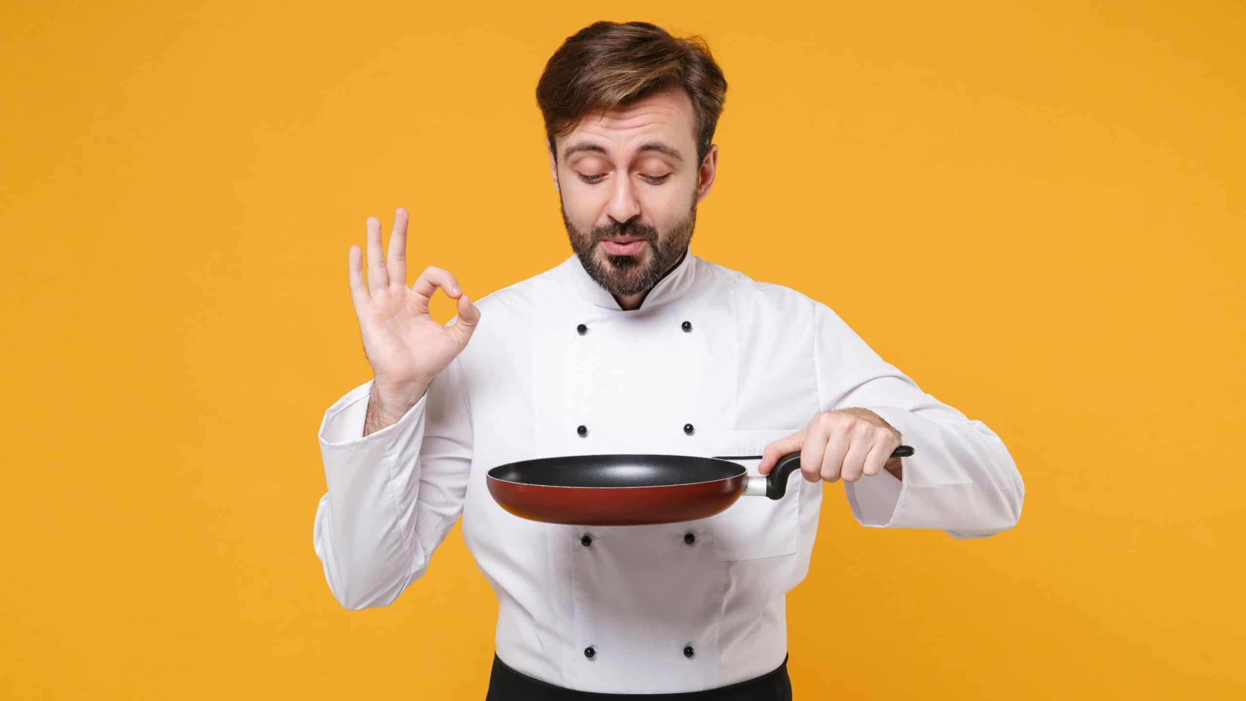 Young bearded male chef cook or baker man in white uniform shirt posing isolated on yellow wall background in studio. Cooking food concept. Mock up copy space. Holding frying pan, showing OK gesture