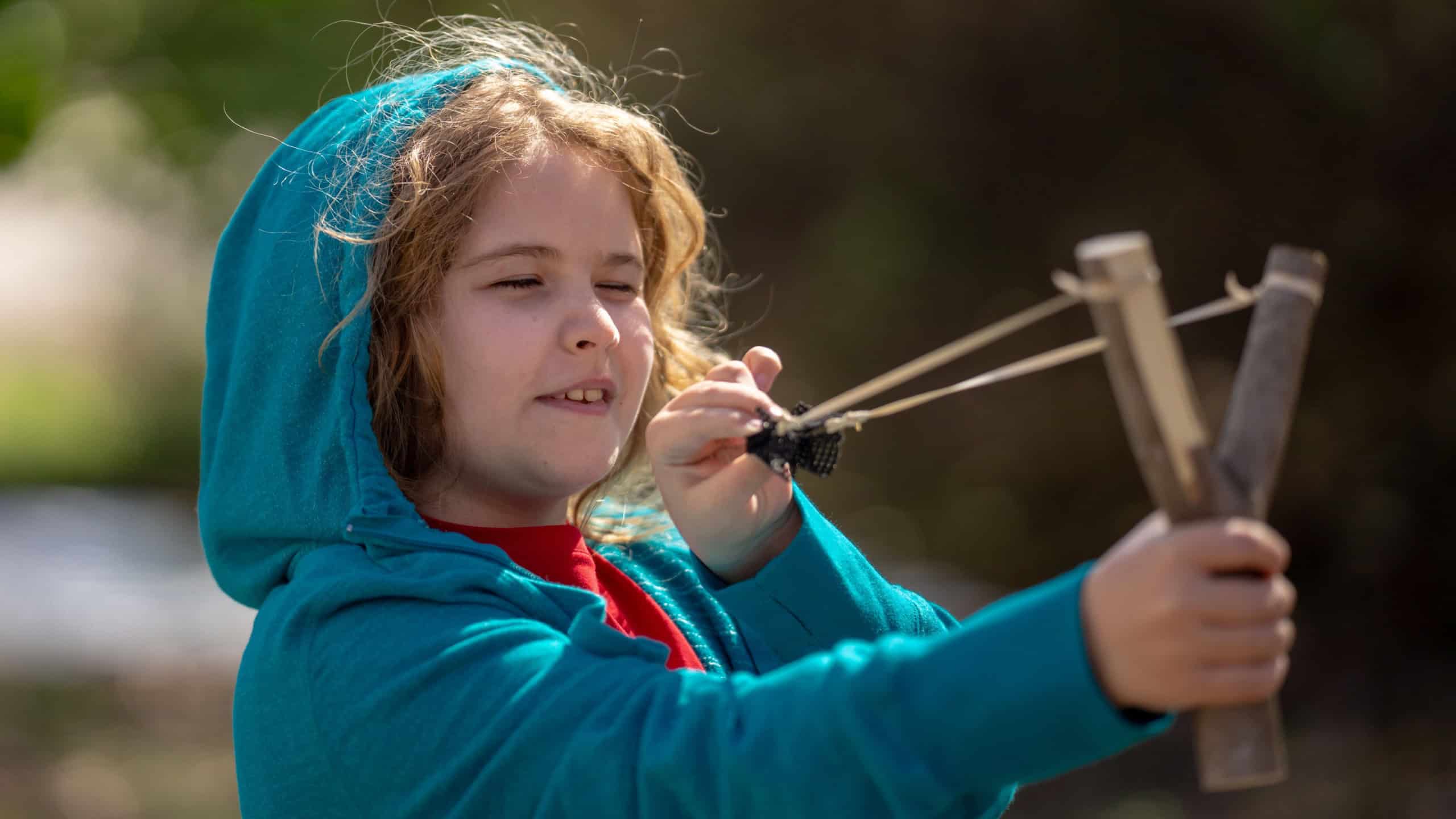 Slingshot in kids hand. Child holding catapult. Boy aiming with sling. Child playing with slingshot. Handmade wooden slingshot. Child aiming at target. Child with slingshot. Outdoor fun with catapult.