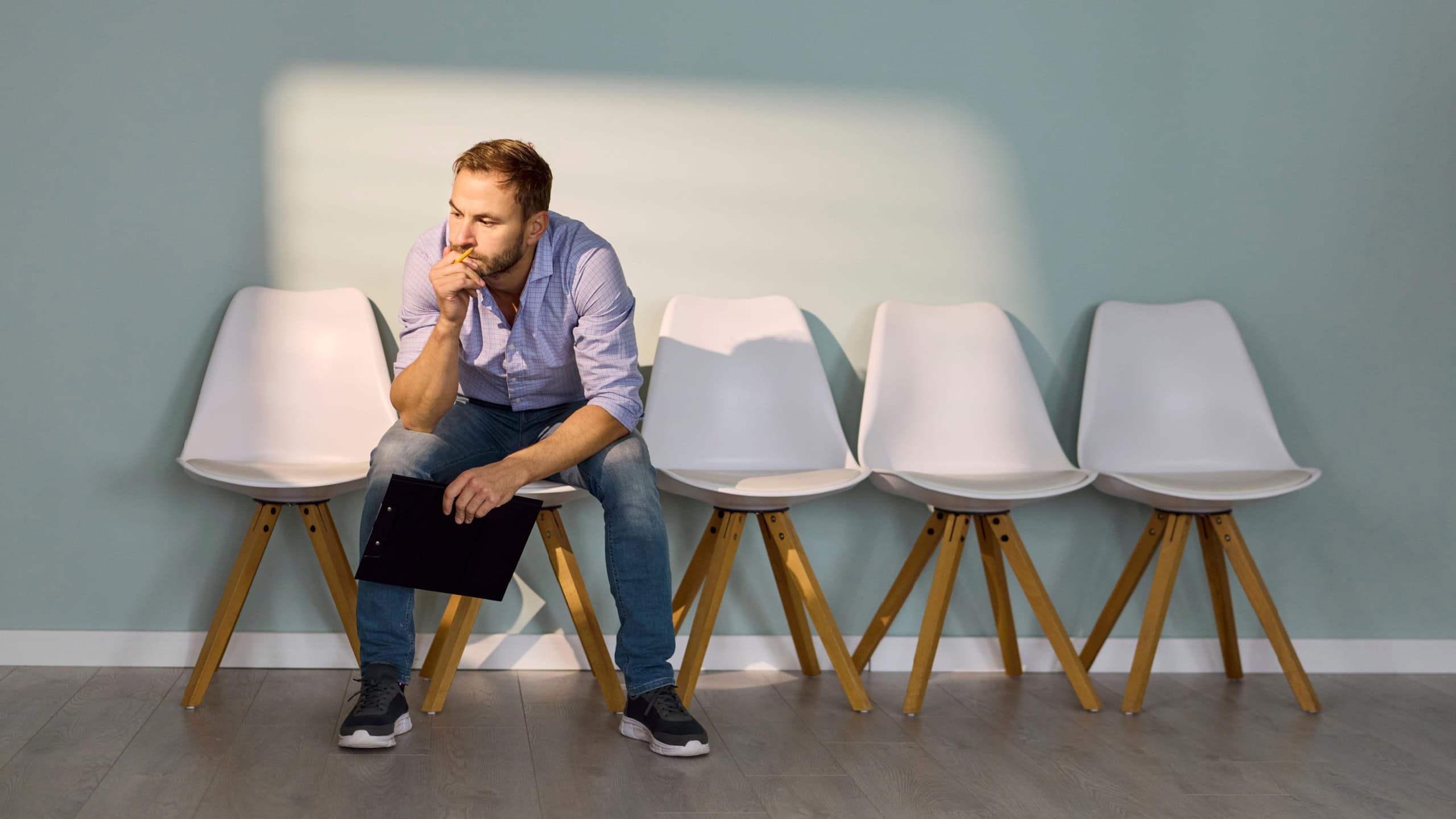 Man employee waiting for interview in office corridor. Male job seeker and candidate sits on chair with a resume, rehearsing answers as anxiety grows in the quiet lobby. Recruitment concept