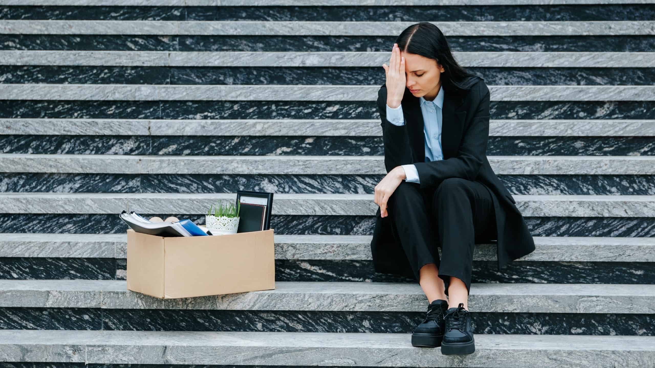 With a heavy heart, the woman sits on stairs, processing her loss of work and unemployment. Unhappy Woman Holding Cardboard Box on Stairs