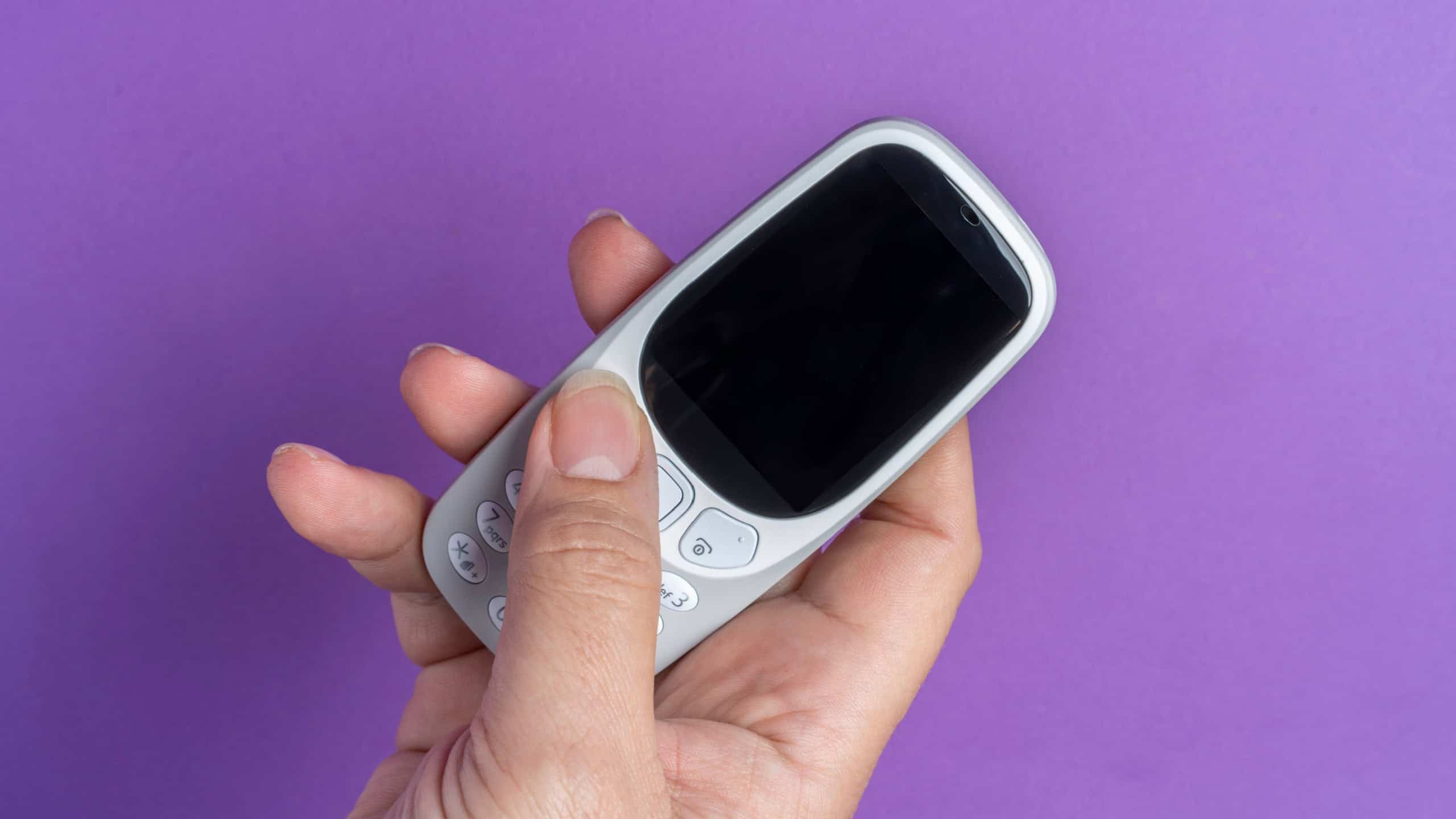 Close-up of a hand holding a classic push-button mobile phone on a purple backdrop, illustrating the concept of digital detox and retro technology.