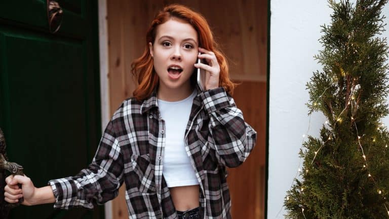 Image of shocked young woman with red hair open the door. Look at camera while talking by phone.