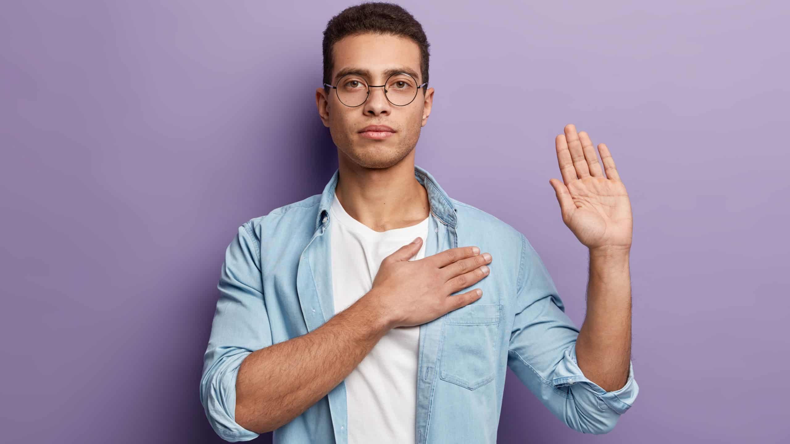 Honest serious hipster guy swears to do something, keeps hand on chest, gestures with palm, promises something, wears spectacles and denim shirt, swears indoor, isolated over purple background