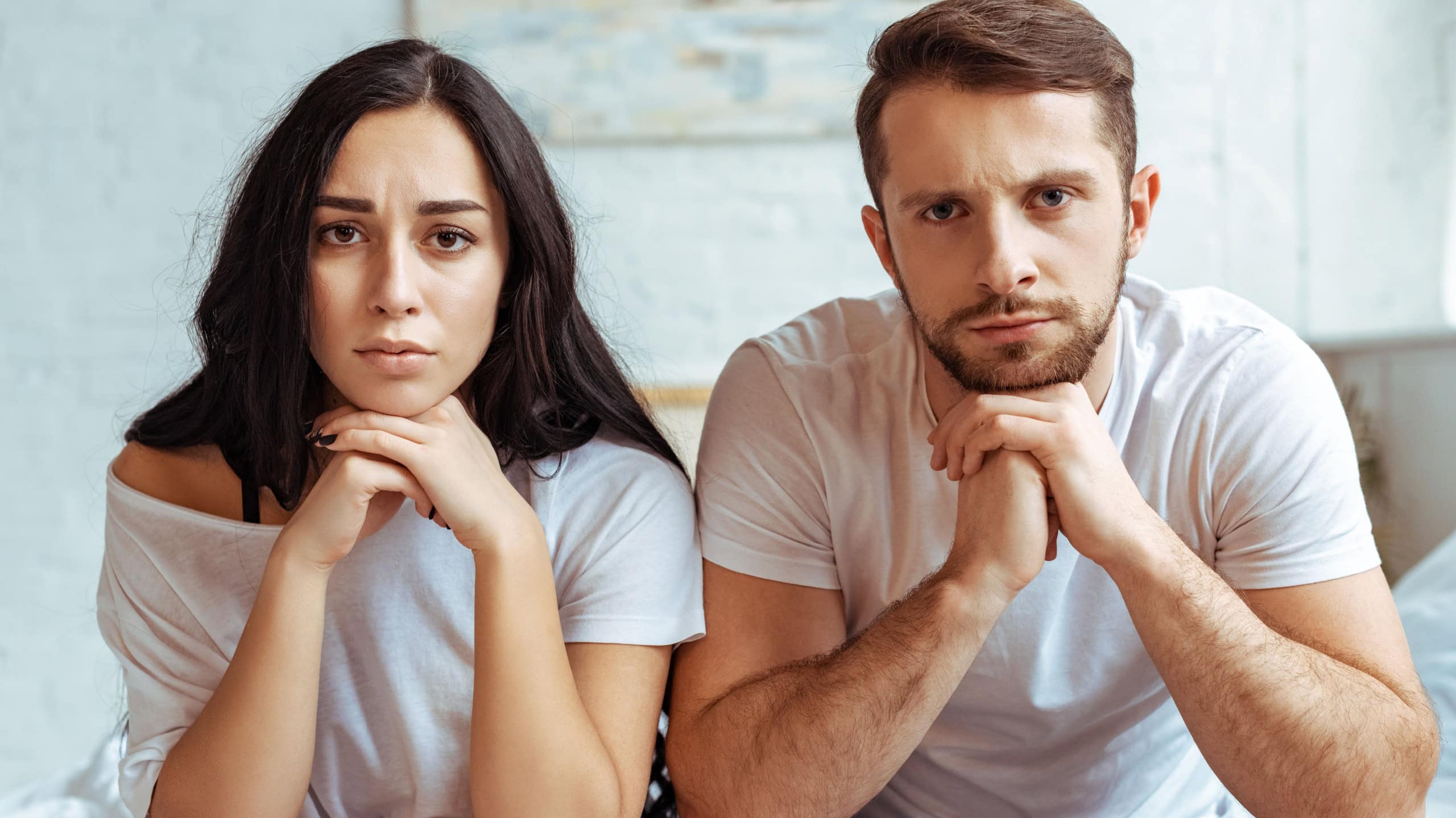 Handsome man in jeans and beautiful sad woman in t-shirt sitting on bed and looking at camera