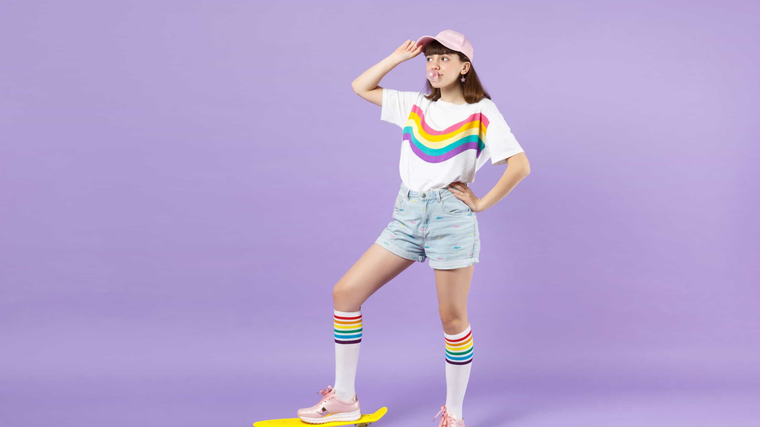 Cute teen girl in vivid clothes standing with yellow skateboard, blowing bubble gum, looking aside isolated on violet pastel background. People sincere emotions, lifestyle concept. Mock up copy space
