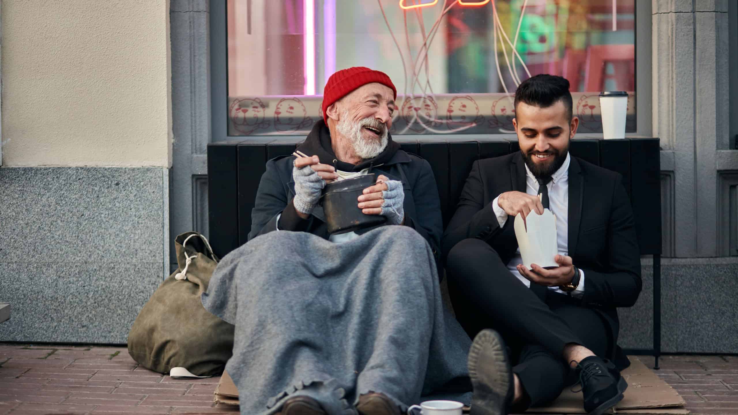 Stylish office worker and beggar male sit eating on street and speaking. Man un tuxedo, poor male in old clothes
