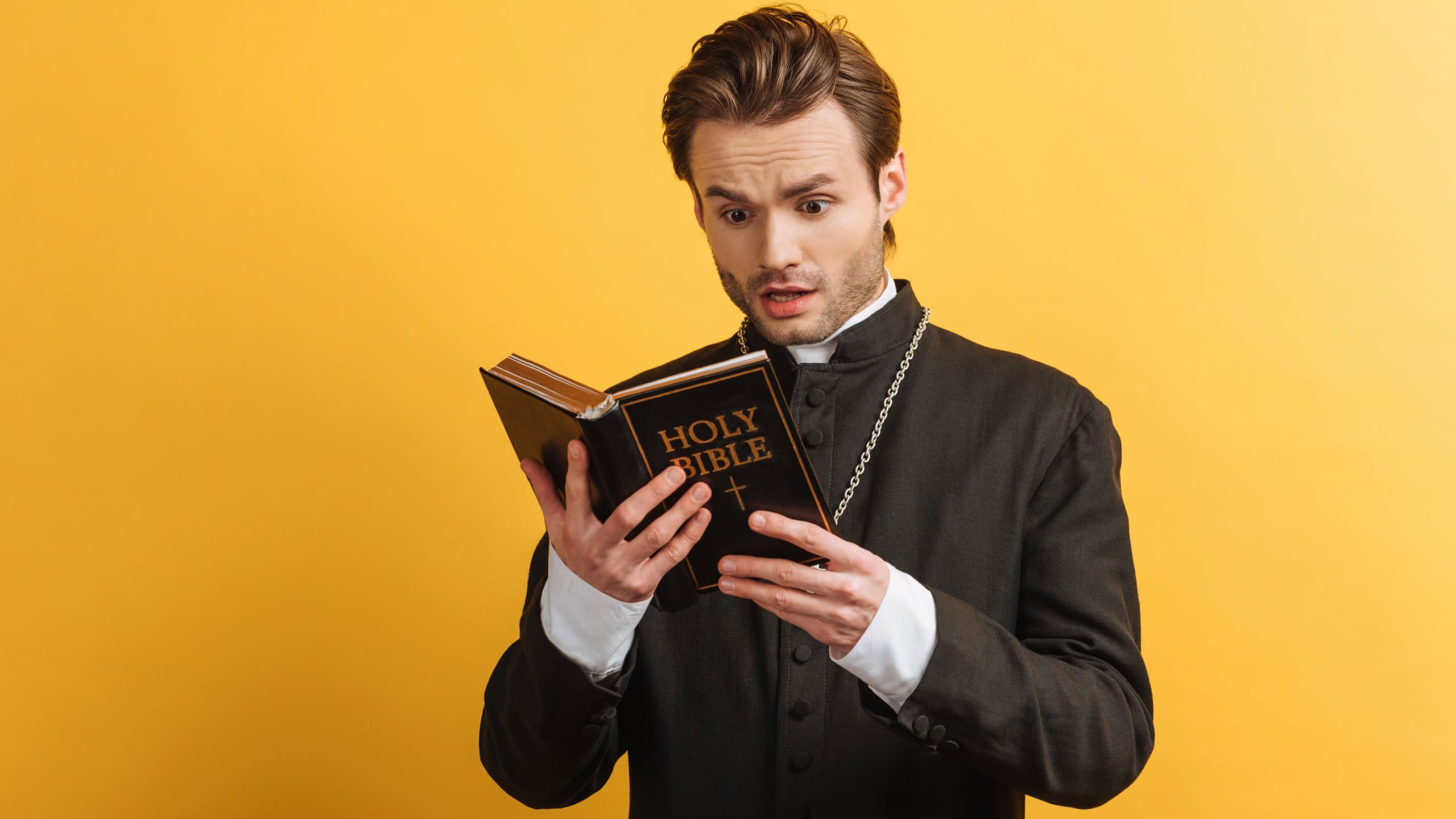 Shocked catholic priest reading holy bible isolated on yellow