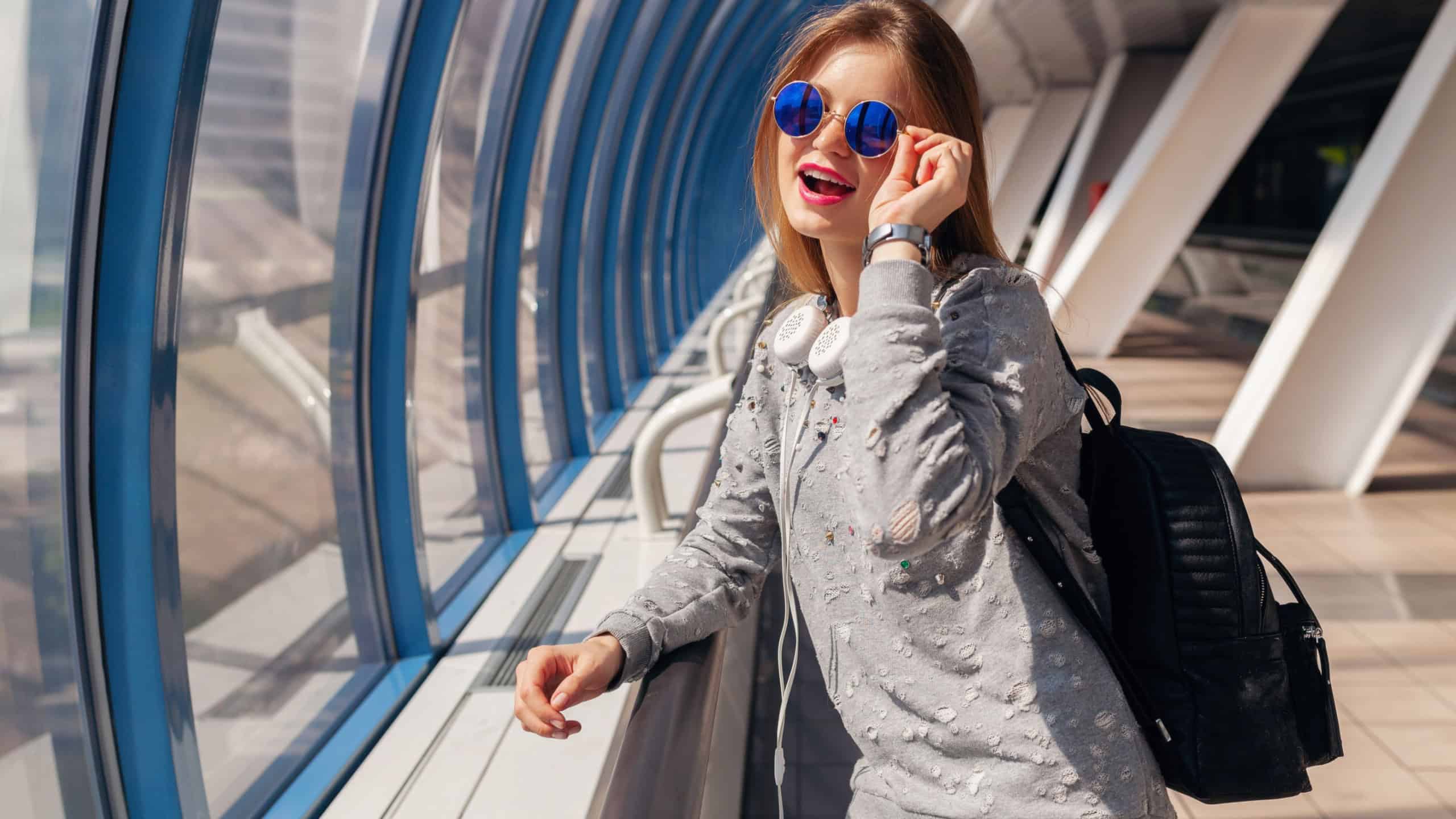 young hipster woman with backpack in urban modern building dressed in casual outfit, student on vacation, wearing jeans, sweater and sunglasses