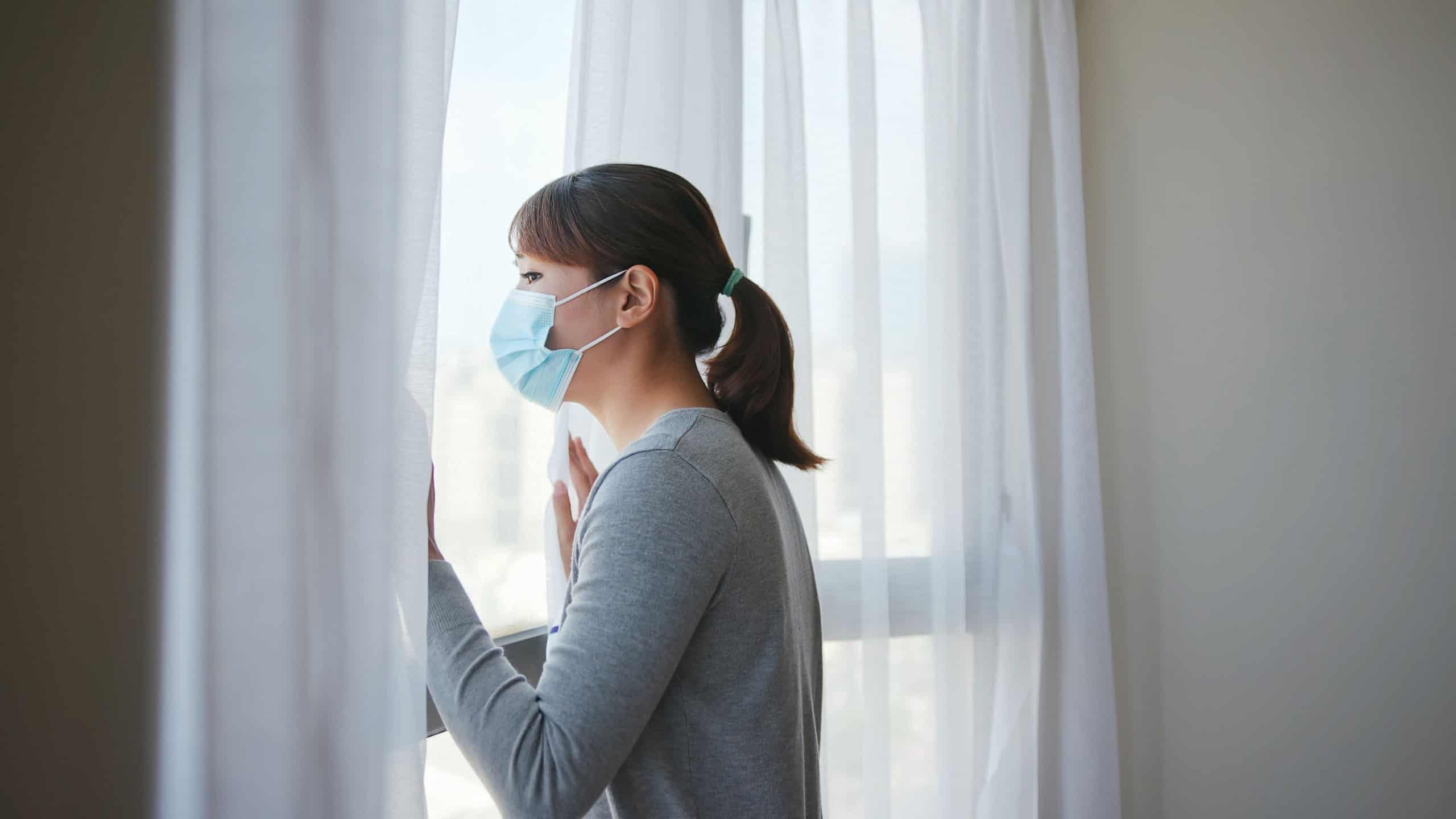 side view asian young woman wearing face mask stay isolation at home for self quarantine due to epidemic of COVID19 - she looks out the window