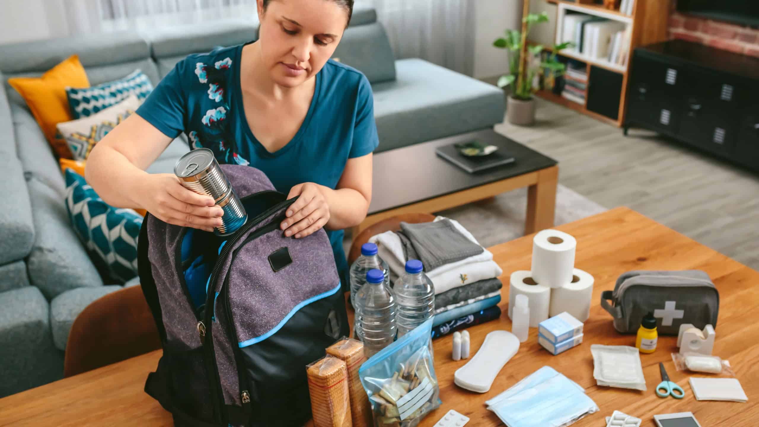 Woman putting cans of food to prepare emergency backpack in living room
