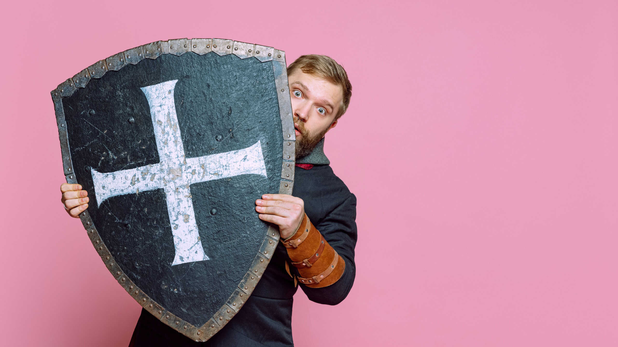 Portrait of scary man, medieval warrior or archer in armor hiding himself behind a shield isolated over pink studio background. Frightened fighter. Comparison of eras, history, renaissance style
