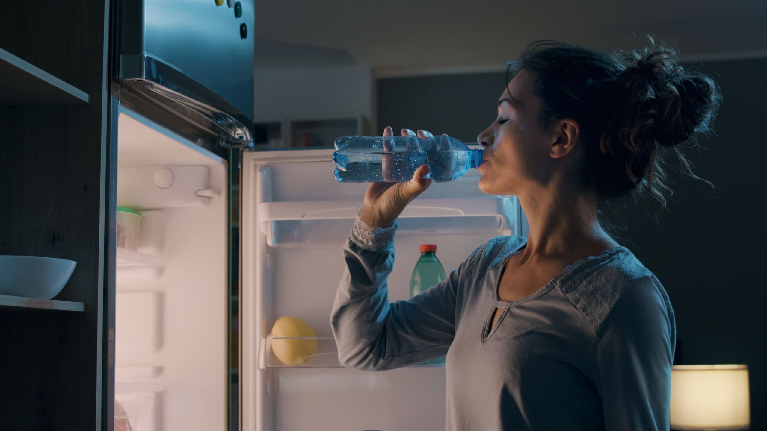 Thirsty woman standing in front of the fridge and drinking water late at night