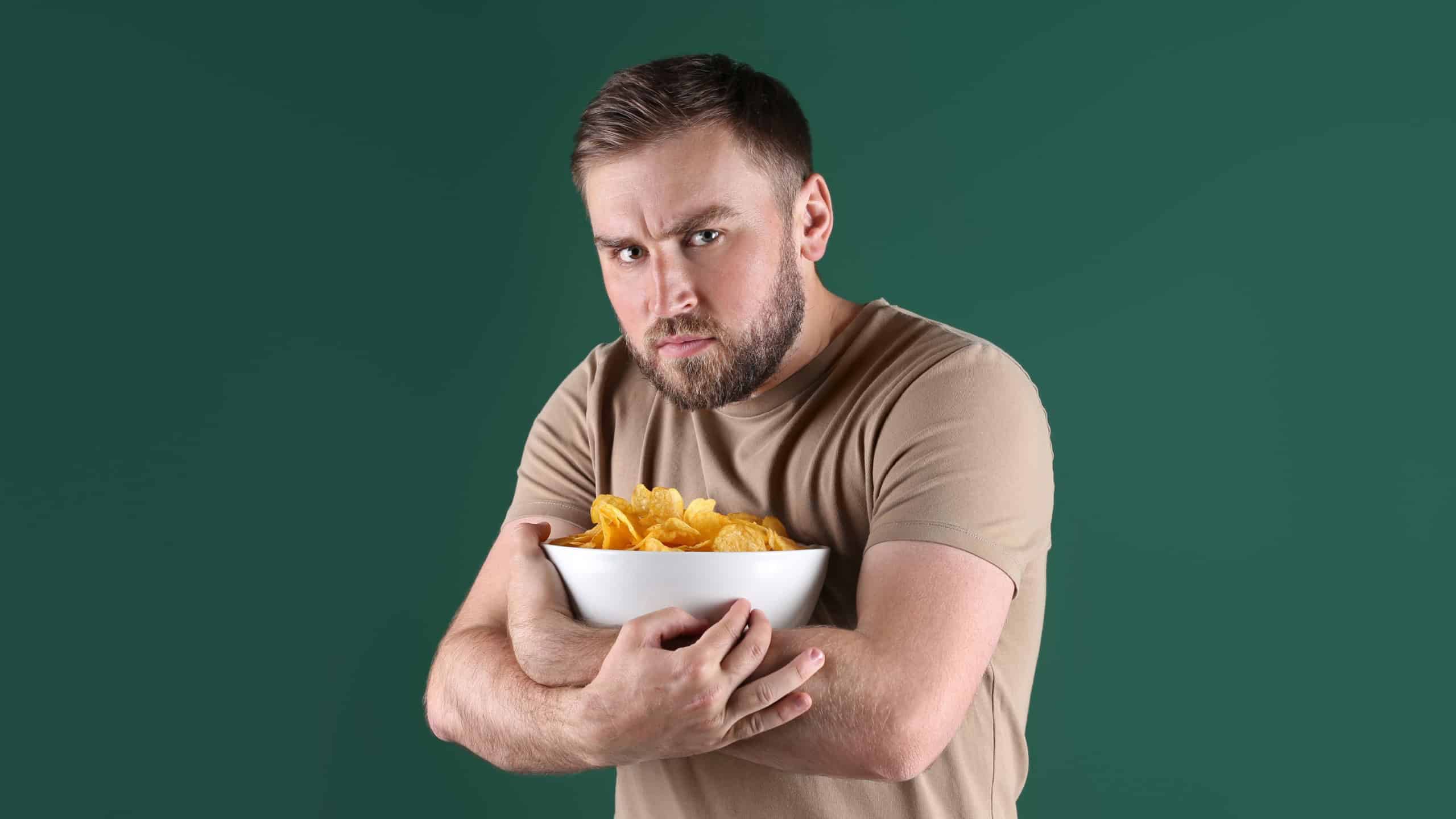 Greedy young man hiding bowl with chips on green background