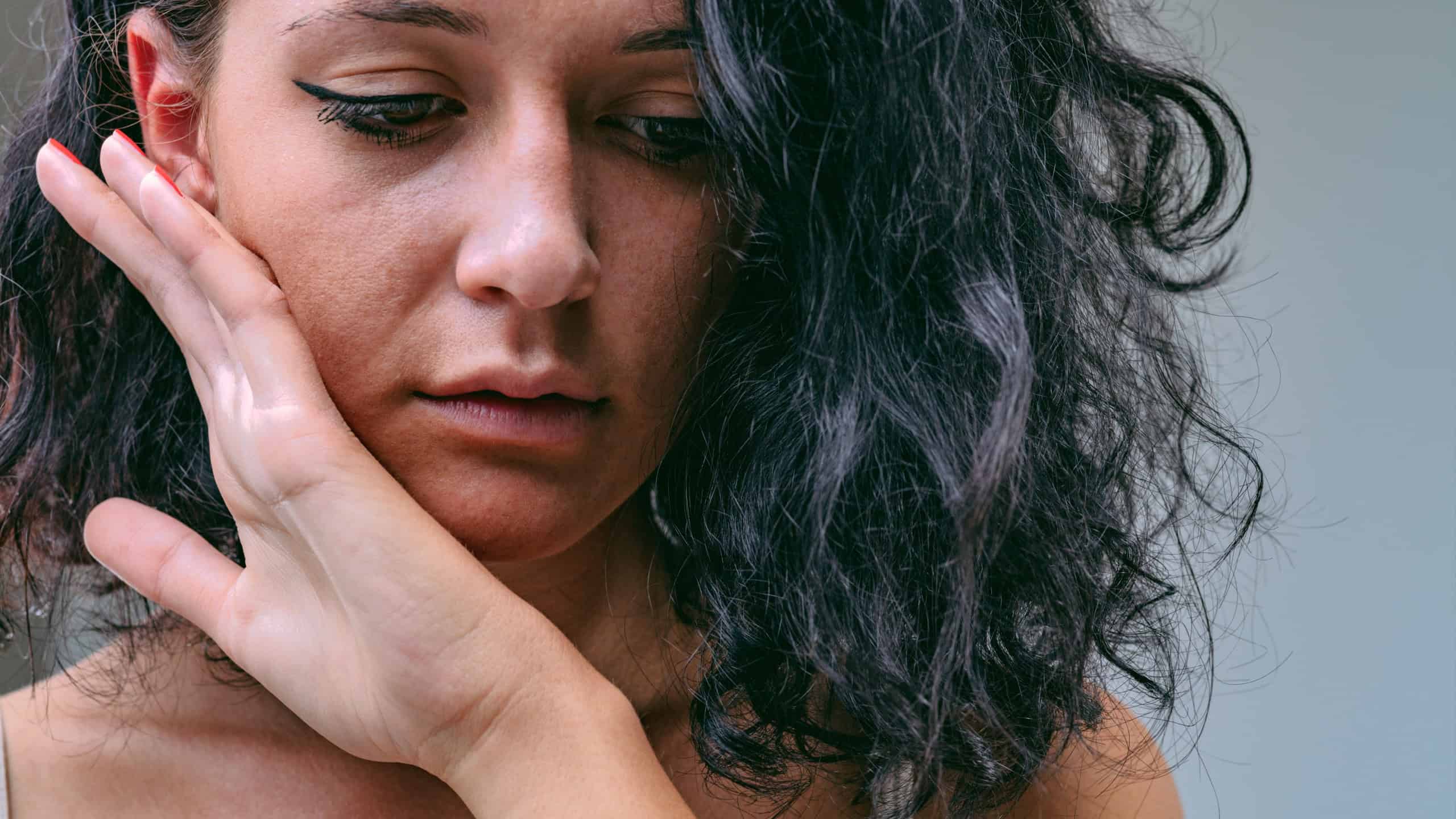 Frontal close-up portrait of young woman bringing her hand to touch the skin of her cheek and chin with the back. She may be feeling how smooth it is, but she may be sad and afraid of a slap.