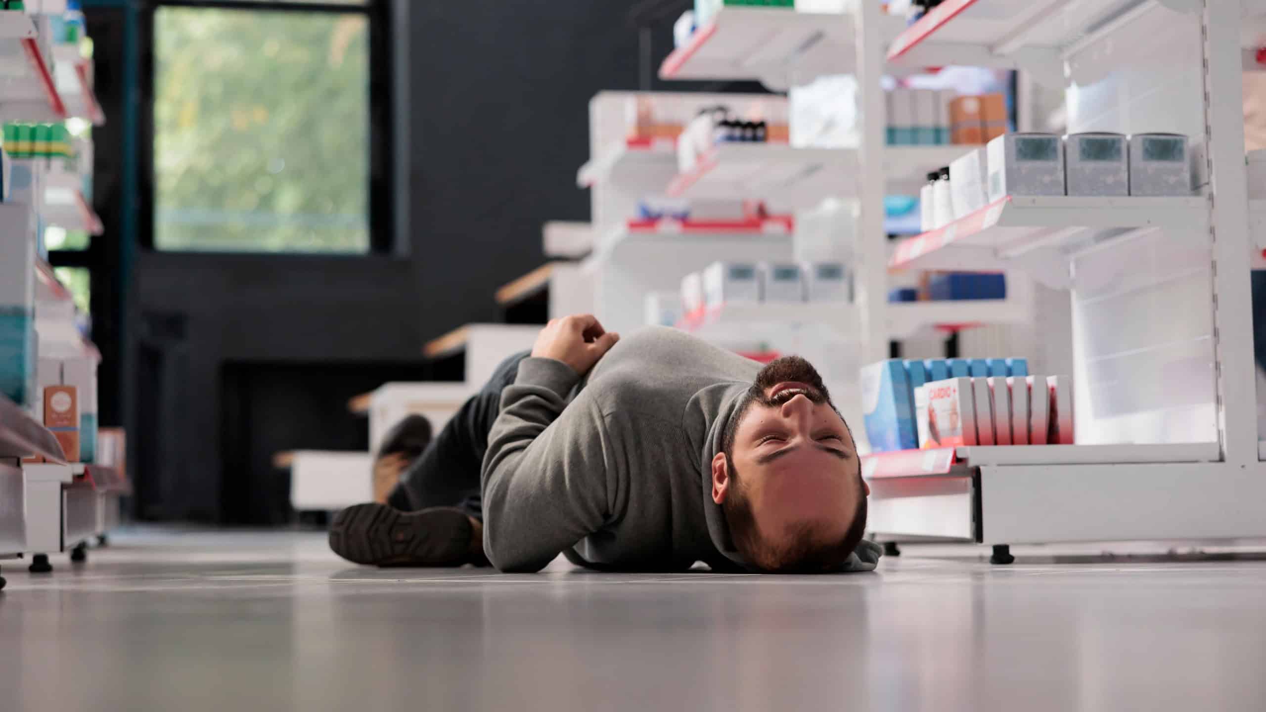 Young caucasian man having epilepsy attack, lying unconscious on floor in medical retail store. Pharmacy store customer fainting, suffering from neurological disease seizure