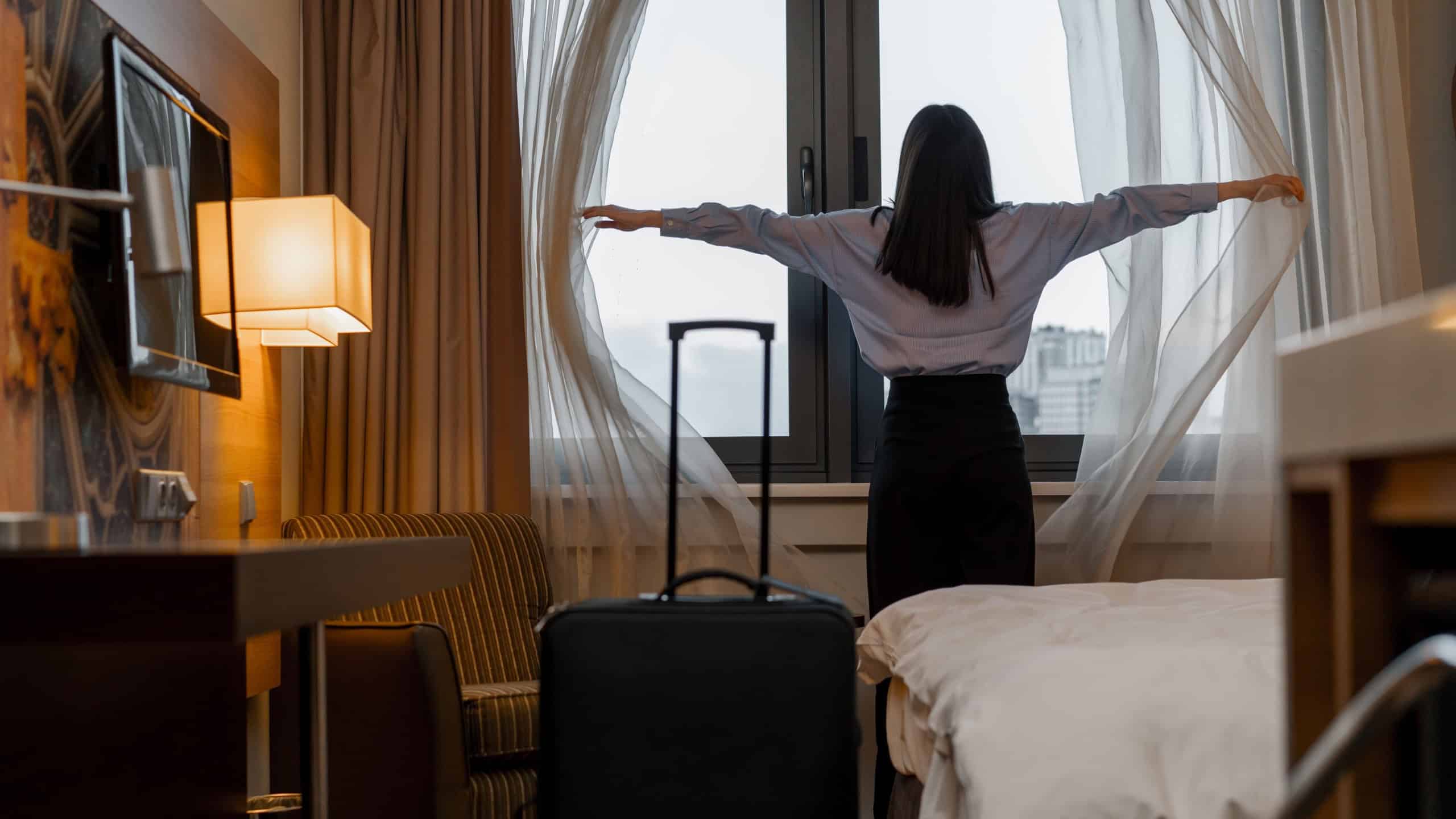 young woman opening curtain while standing luxury hotel room looking through window enjoying city skyscrapers close up