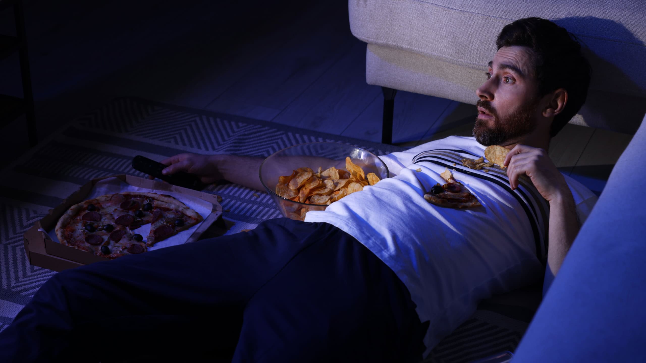 Man eating chips and pizza while watching TV in room at night. Bad habit