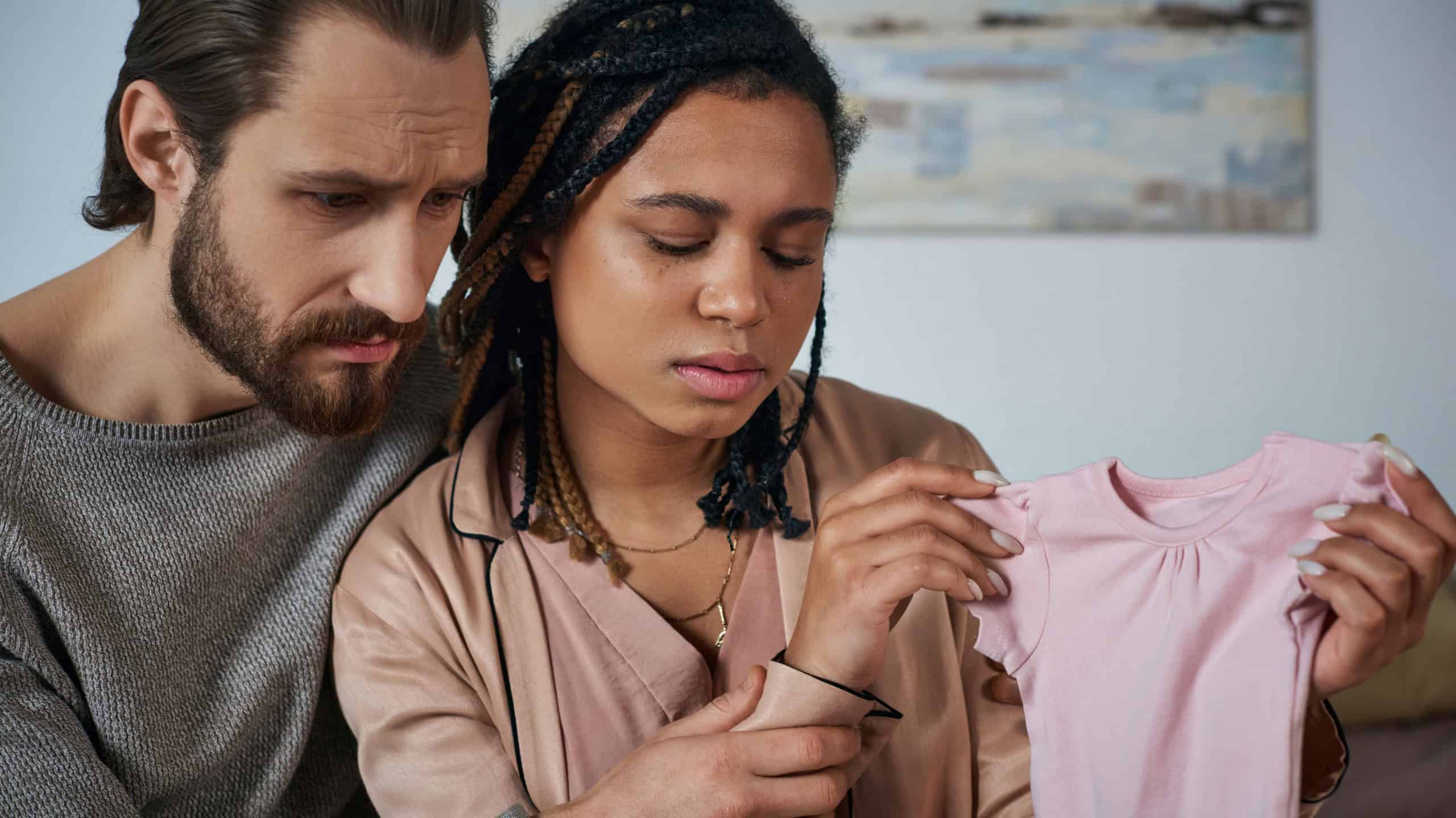 Sad man calming down african american wife crying and holding baby clothes, miscarriage concept