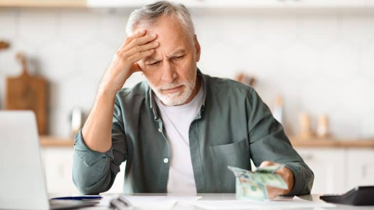 Financial Problems. Depressed Senior Man Looking At Cash Money In Hand While Sitting At Table In Kitchen, Upset Older Gentleman Counting Banknotes, Having Economy Crisis After Retirement