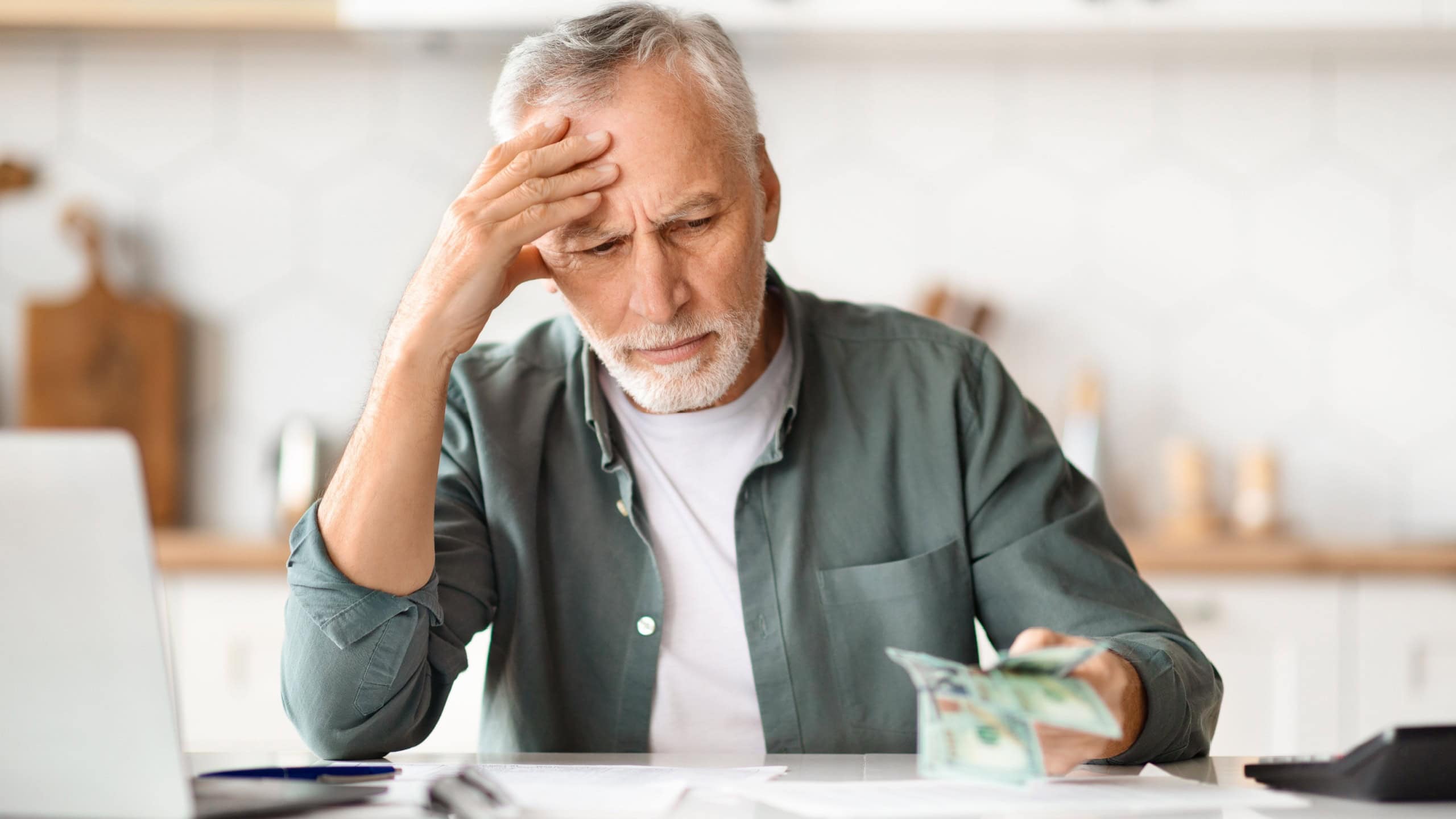 Financial Problems. Depressed Senior Man Looking At Cash Money In Hand While Sitting At Table In Kitchen, Upset Older Gentleman Counting Banknotes, Having Economy Crisis After Retirement