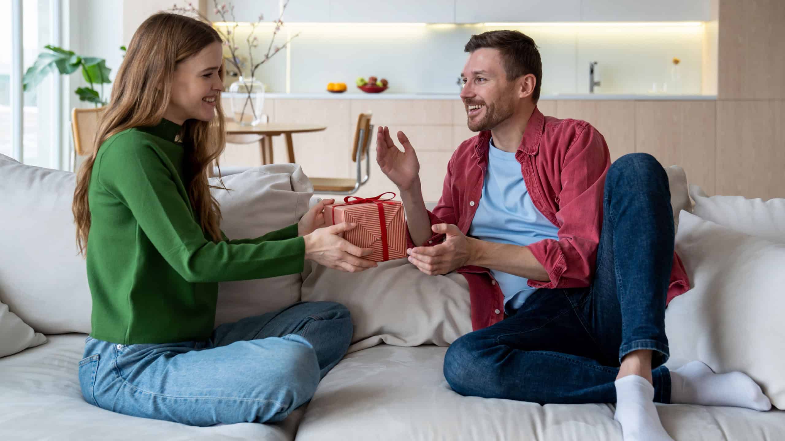 Pleased wife congratulating husband giving present gift box sitting on couch at home. Happy couple celebrating birthday, anniversary, family holiday event together. Female makes surprise to boyfriend