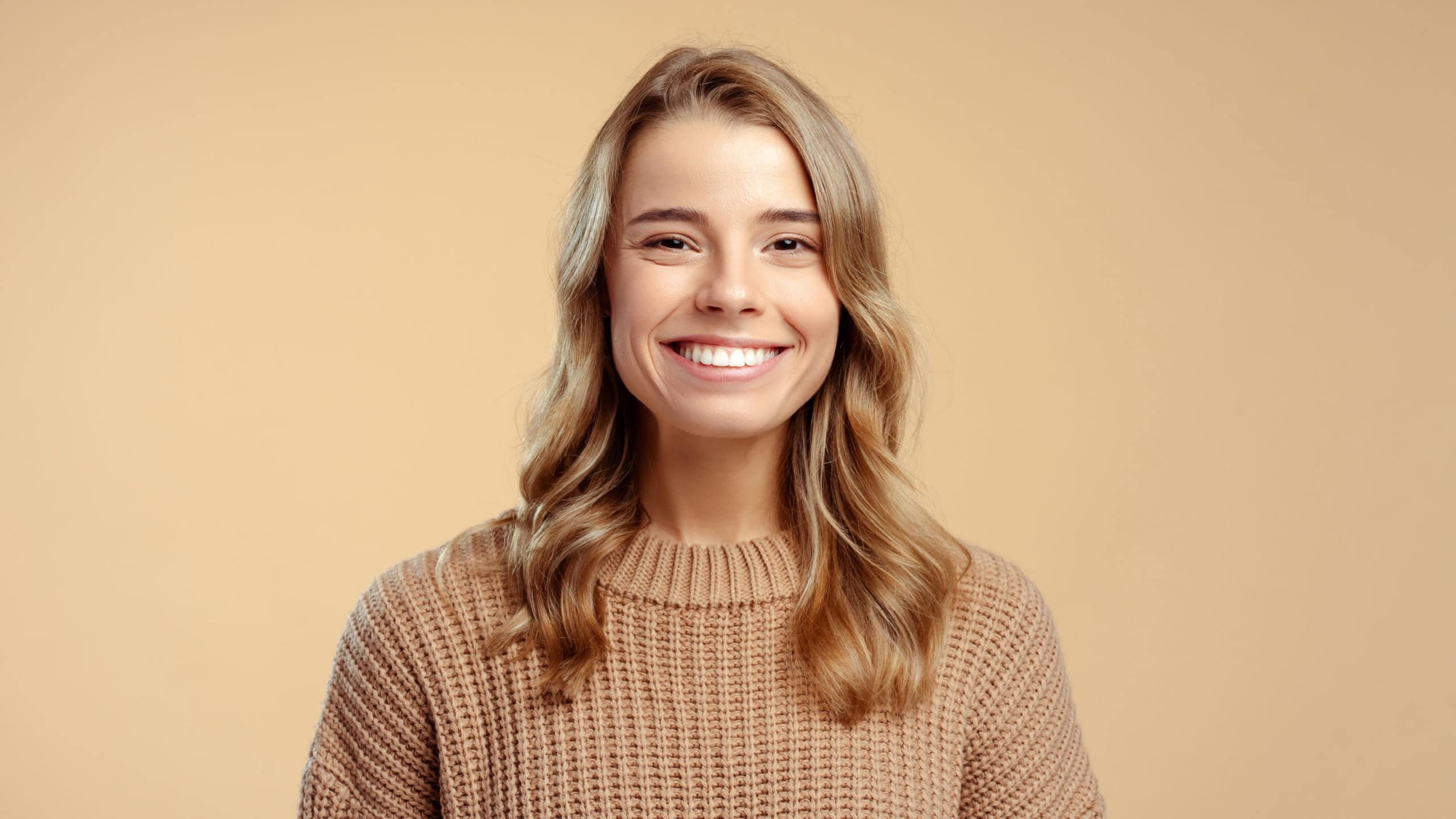 Portrait of smiling attractive young woman wearing stylish warm winter sweater looking at camera standing isolated on beige background. Concept of natural beauty