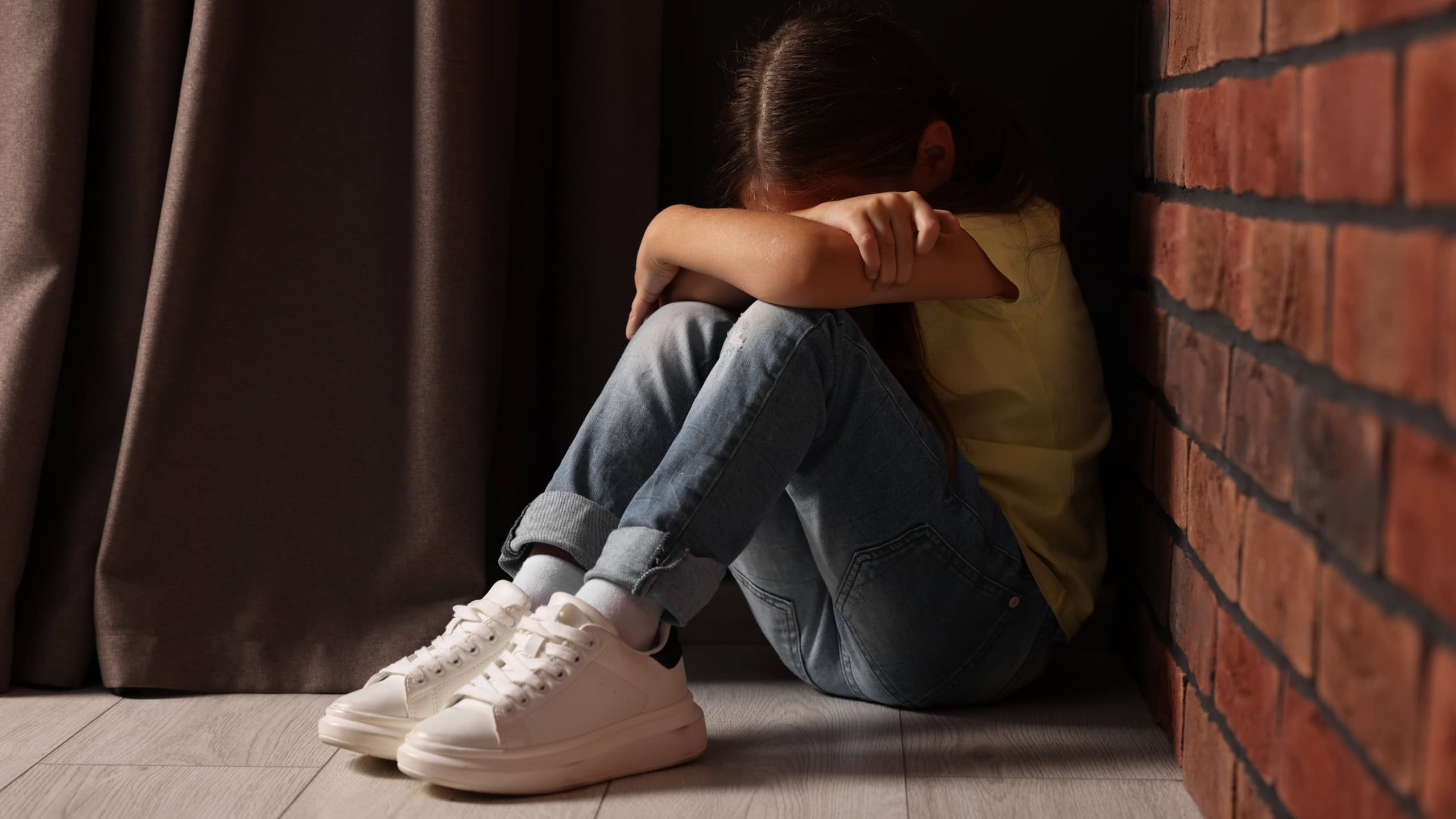 Child abuse. Upset little girl sitting on floor near brick wall indoors