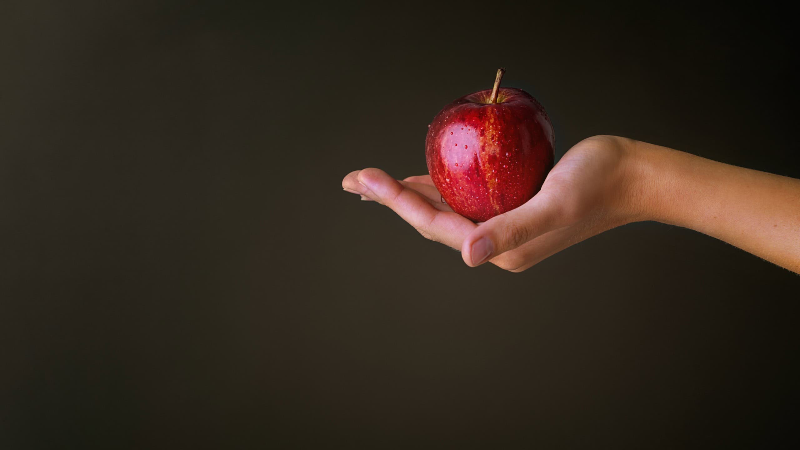Giving, hand and red apple in studio for nutrition, wellness and healthy living with vitamin c for diet. Mockup, organic and forbidden fruit of knowledge, temptation and person by dark background