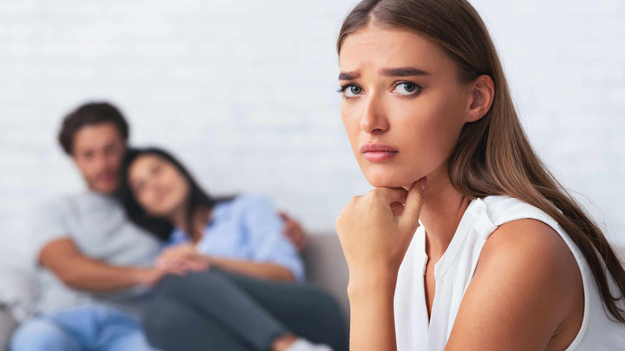 Jealousy. Sad Girl Looking At Camera While Her Ex-Boyfriend Hugging New Girlfriend Sitting On Couch Indoor. Selective Focus