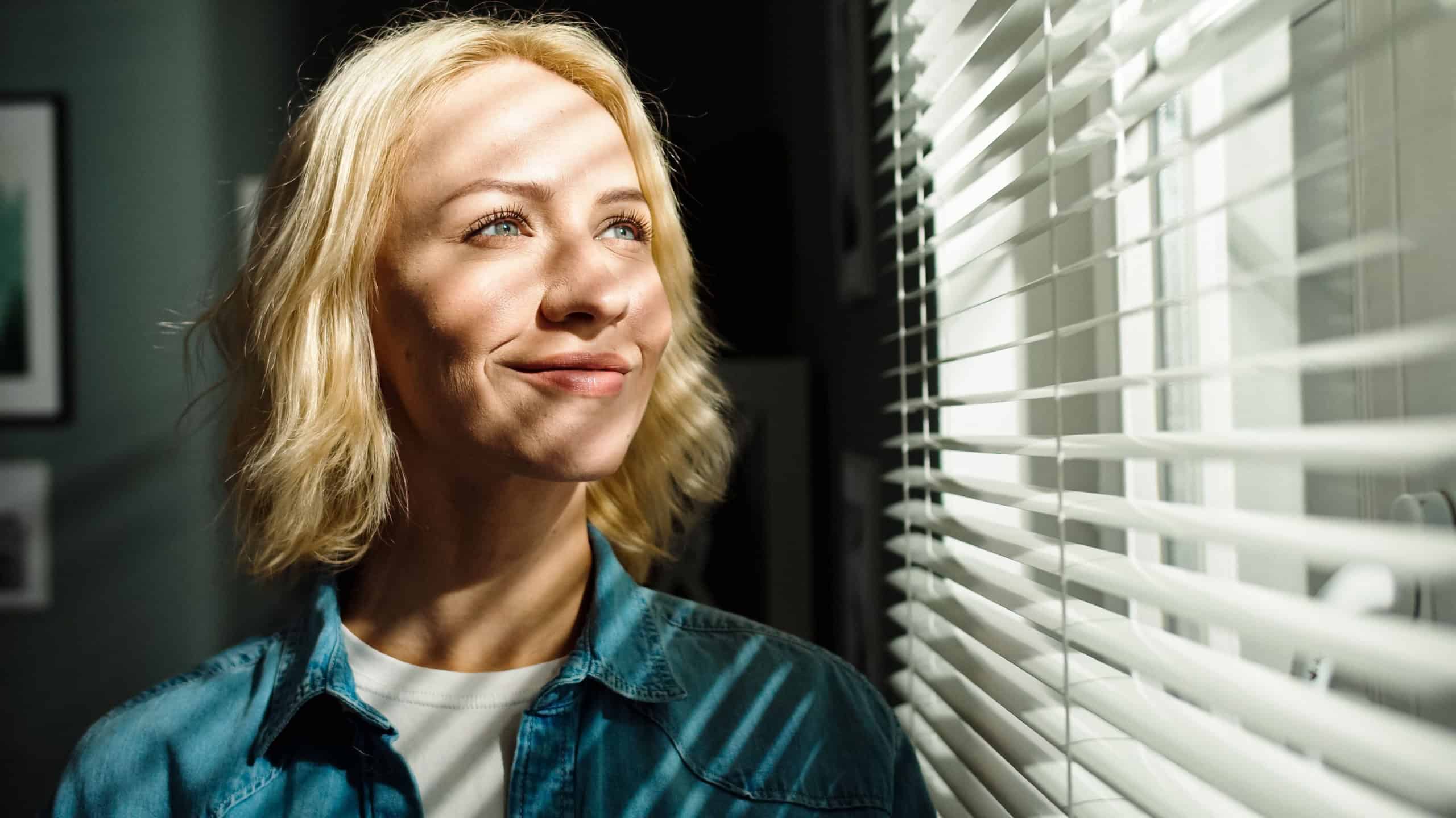 Close up portrait of beautiful young Caucasian female looking out window with smile on face. Sun's rays fall on woman's face with warm.
