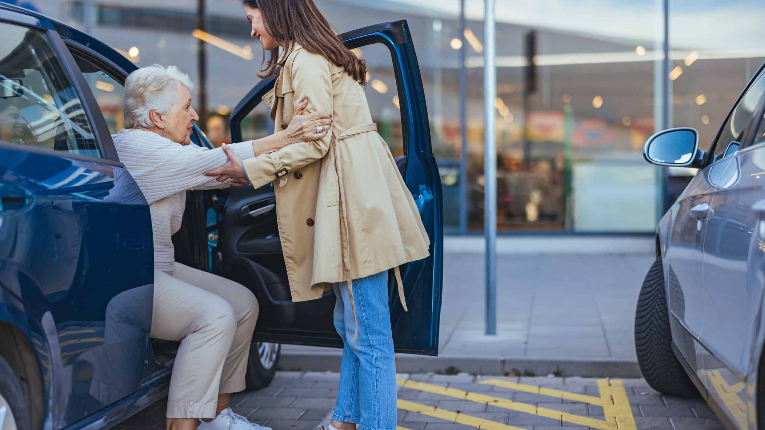 A young woman helps an elderly lady out of a car in a parking lot, showcasing care, support, and kindness. The image highlights intergenerational bonding and assistance.
