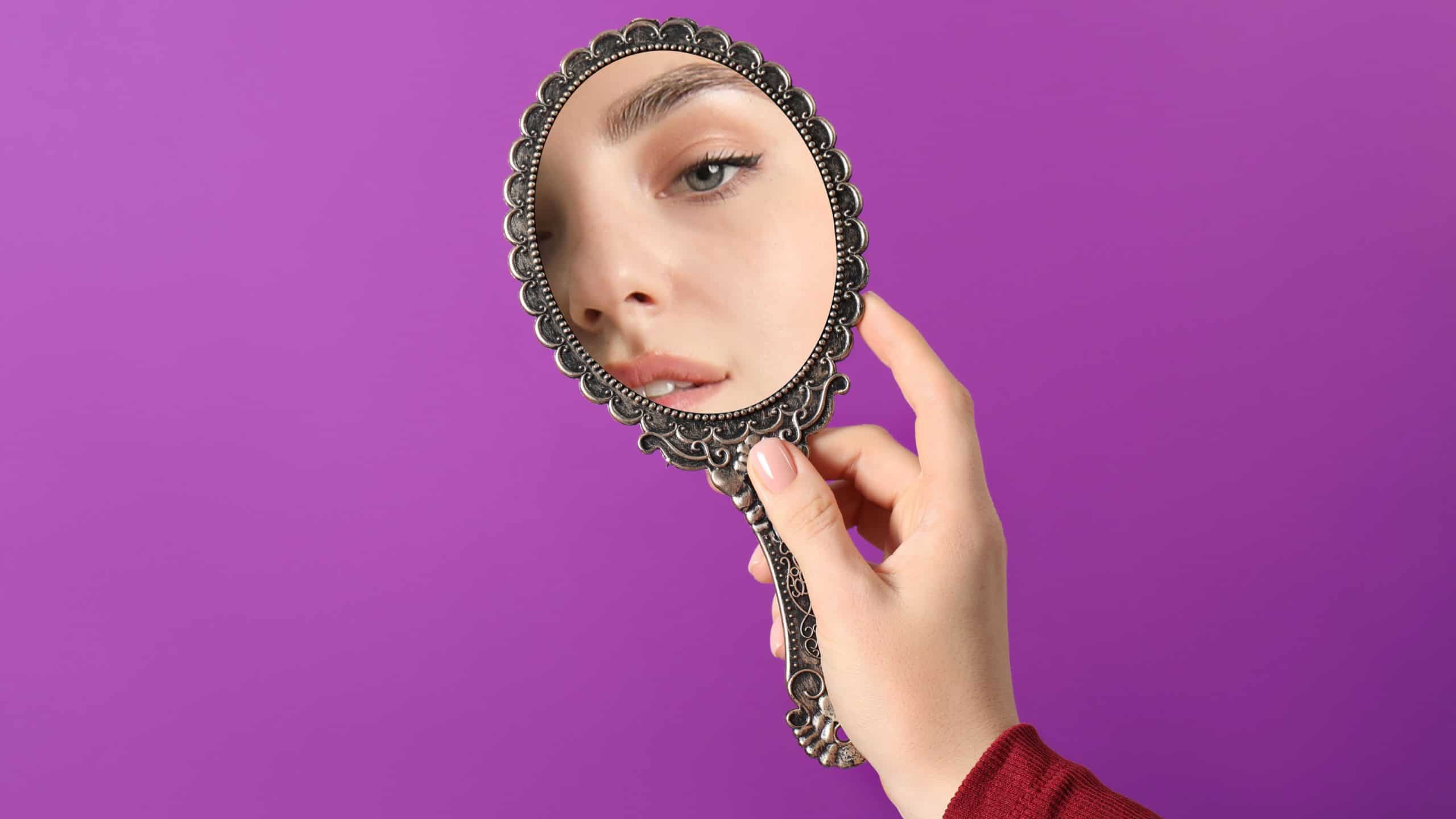 Beautiful woman looking in handheld mirror on purple background, closeup