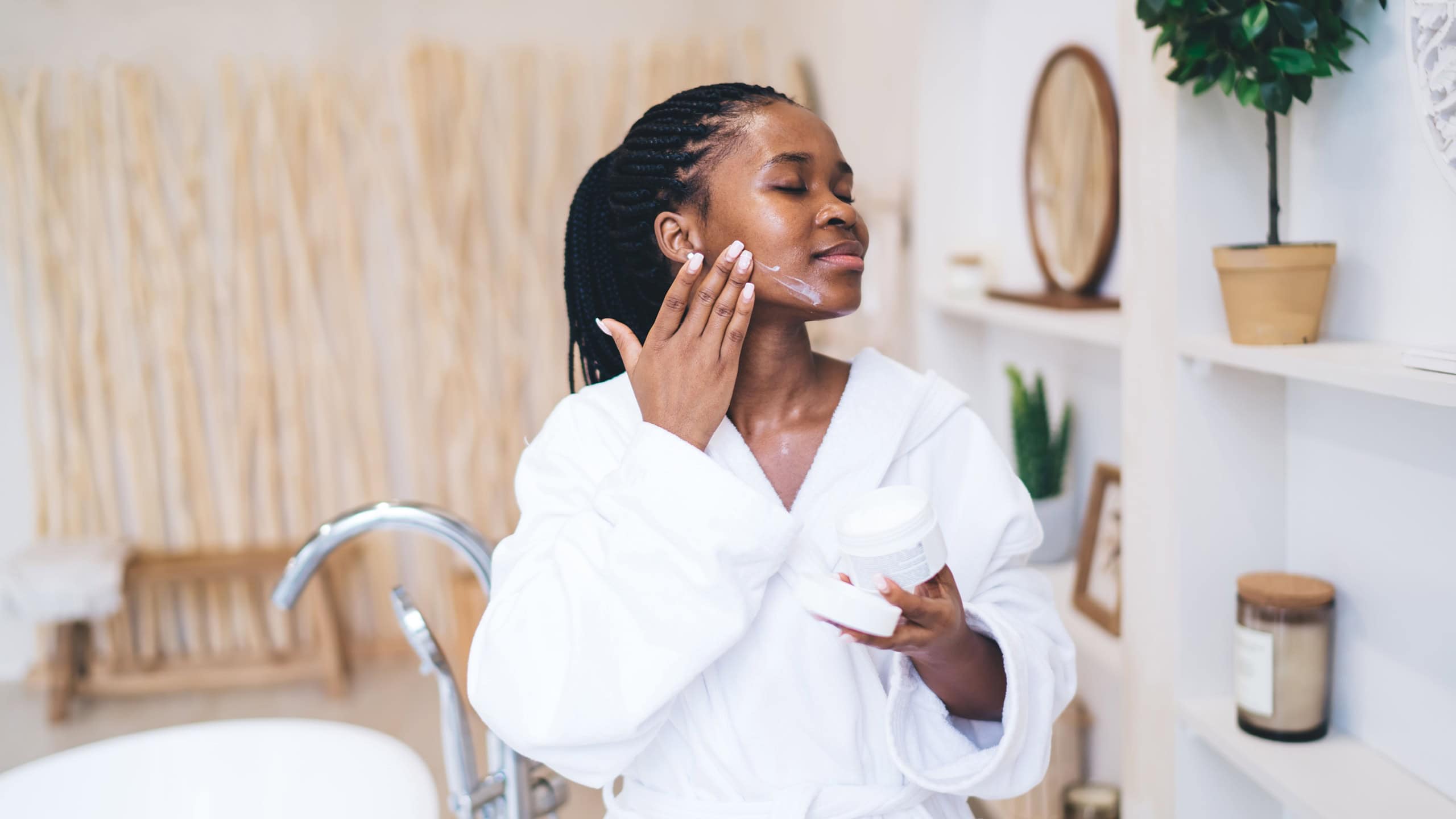 Personal care ritual captured in light bathroom setting - attractive African American woman in robe performing gentle facial selfcare with mindful attention. Charming black female with cream