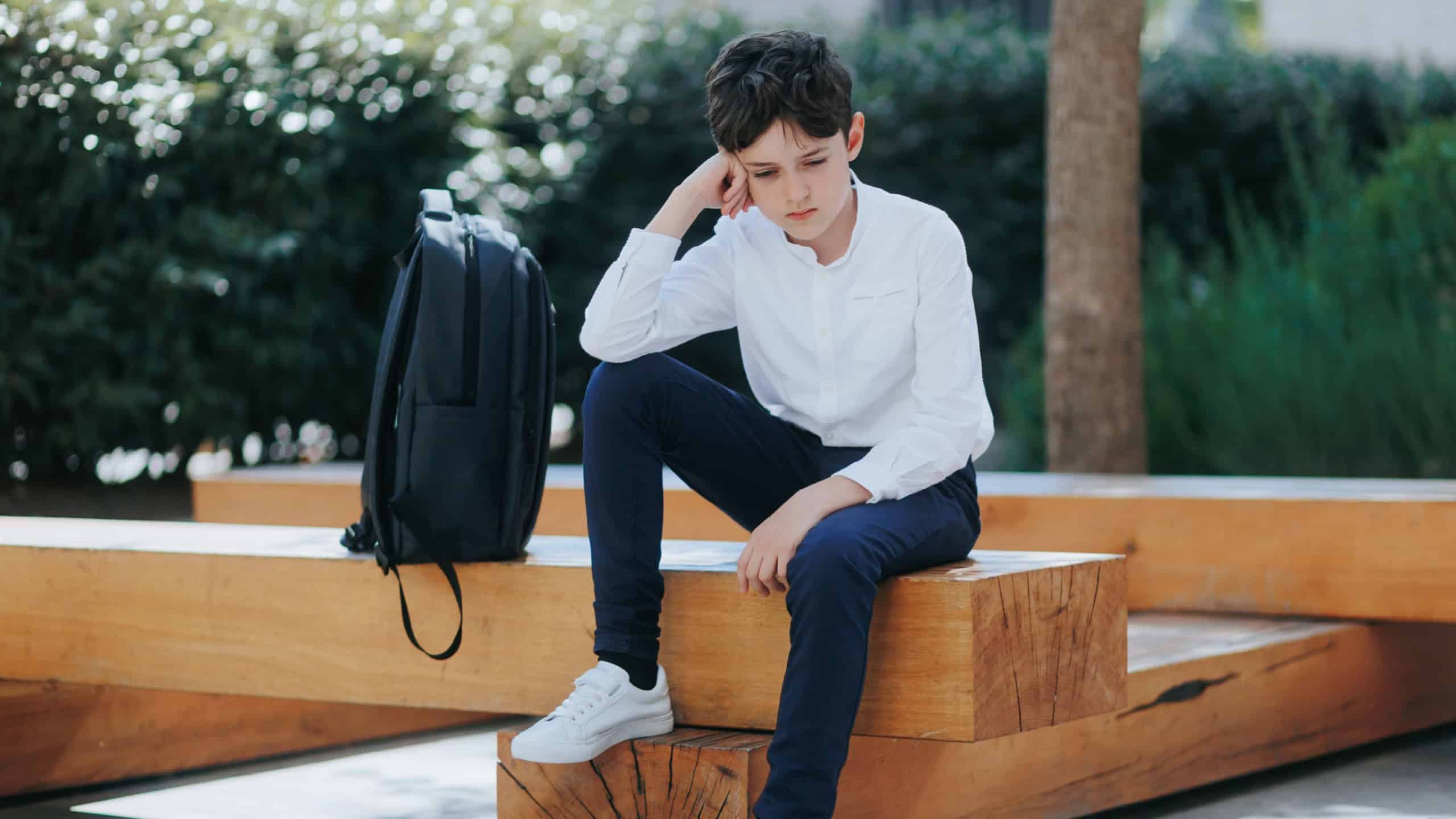 Young schoolboy, about 12 years old, sits silently on a modern bench outdoors. His posture and expression suggest sadness, tiredness, or school-related stress.