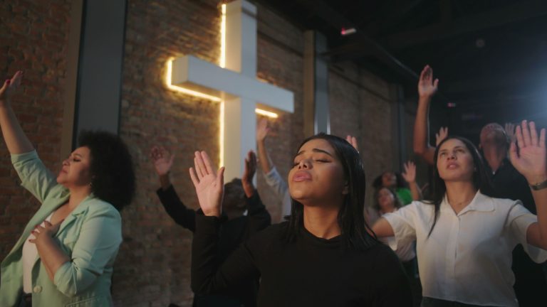 Diverse worshippers raising hands in worship service with glowing cross in background, people deeply involved in prayer and praise