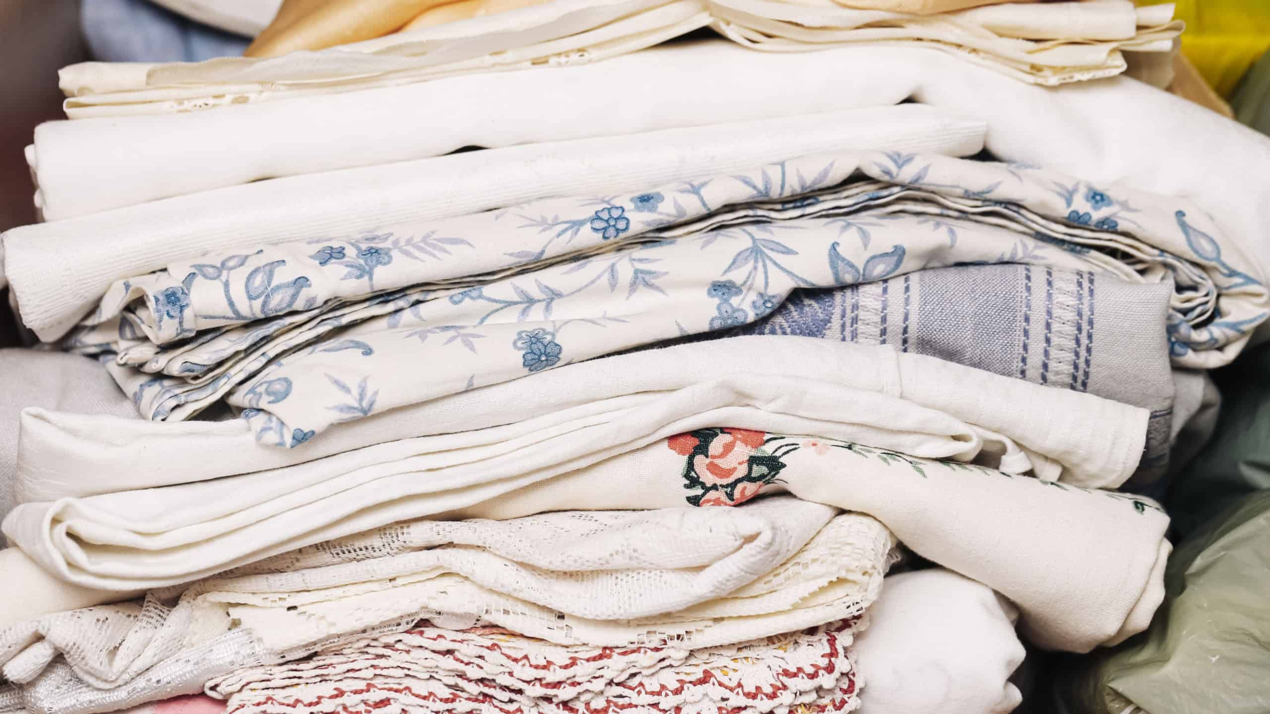 Stack of neatly folded fabric and linens background. Pile of textiles in wardrobe. Mixed patterns and textures. Cotton and lace materials for home use. Organized shelf with clean laundry.