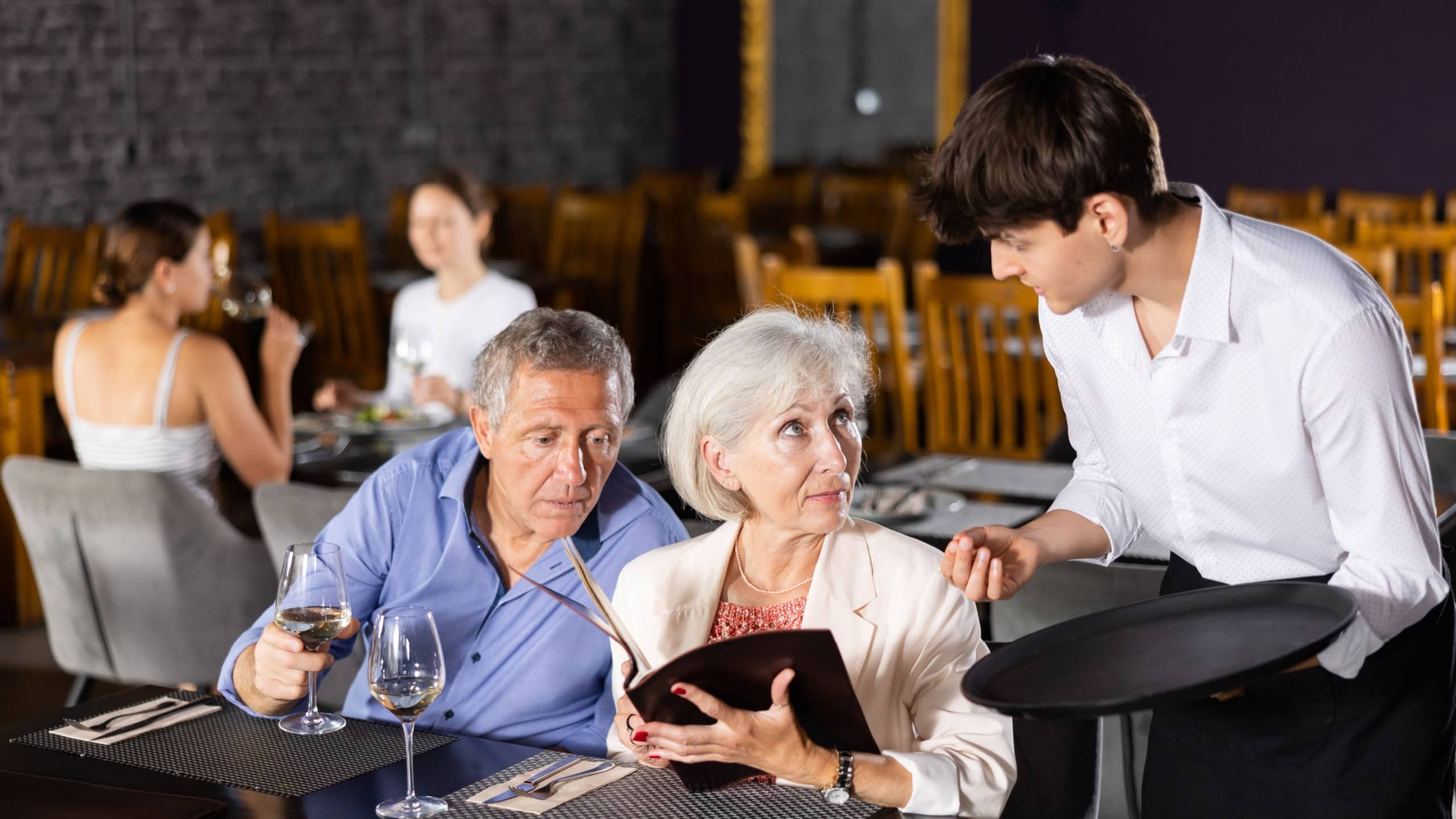 Interested positive senior man and woman sitting at table in restaurant, holding menu and talking to polite young waiter about dishes choice