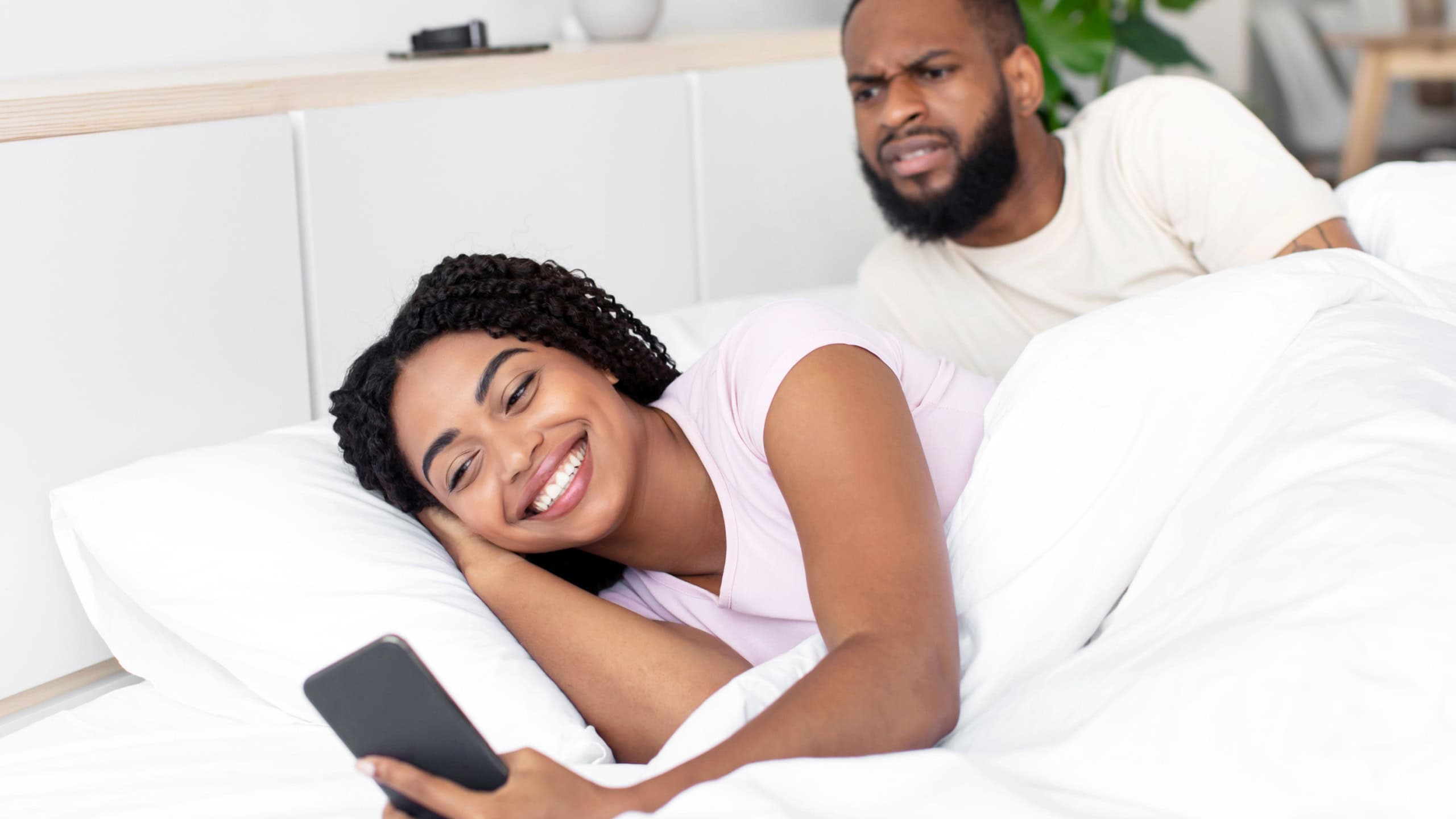 Sad young african american husband looks at wife phone on white bed in bedroom interior