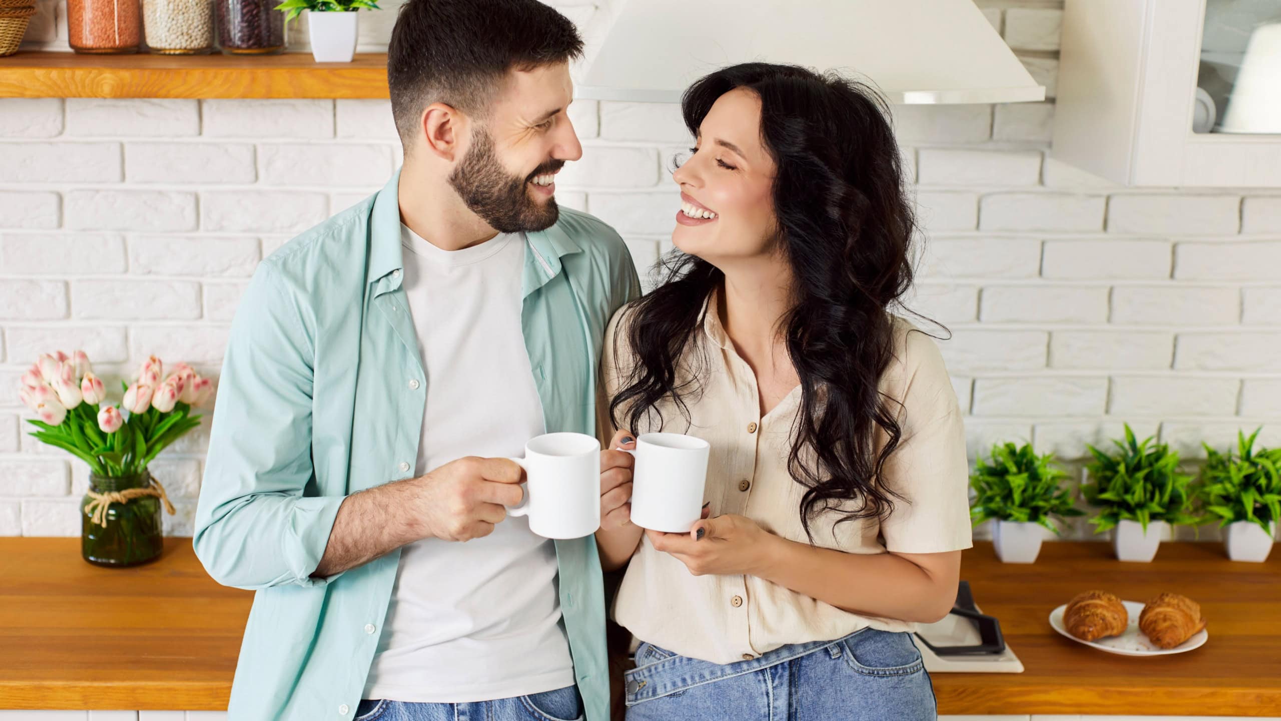 Happy couple enjoying drinking breakfast coffee in cozy home kitchen together. Young people hold mugs and look at each other with love, romantic daily routine of smiling girlfriend and boyfriend