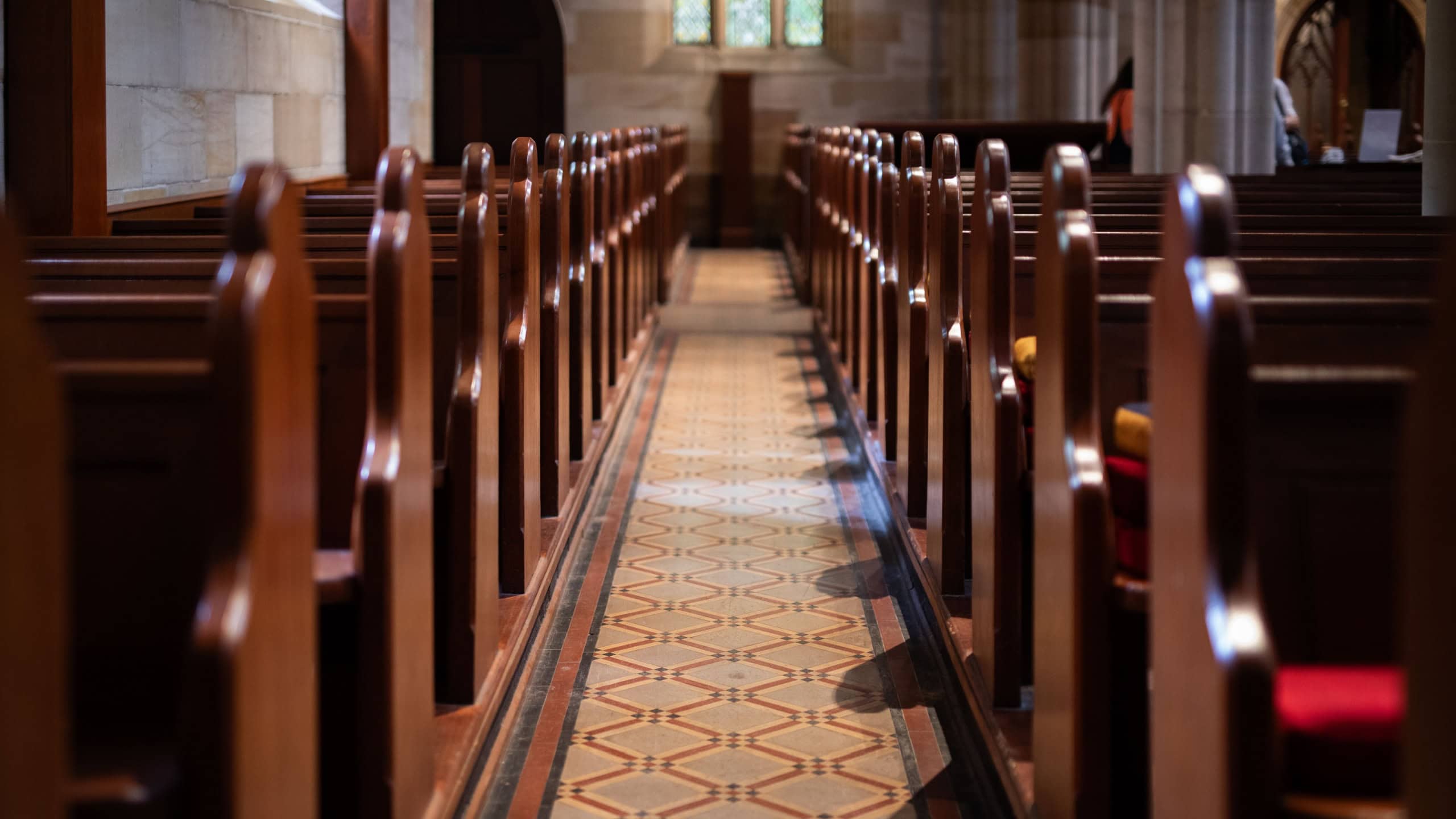 Rows of traditional wooden pews line the patterned aisle inside a quiet, historic church or cathedral.
