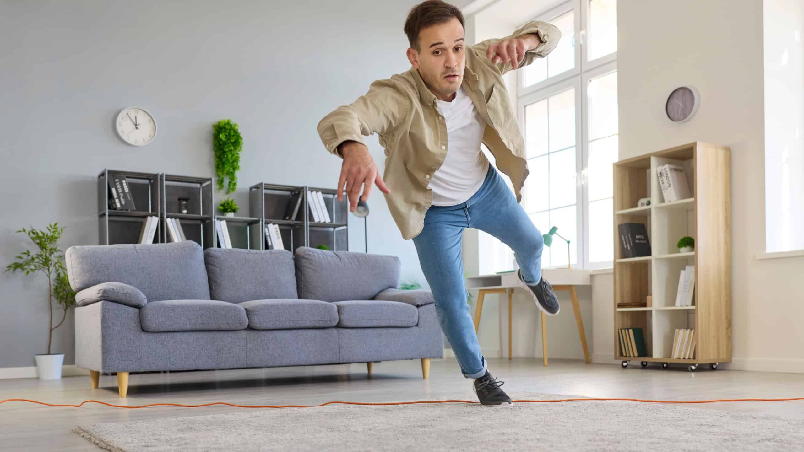 Clumsy man tripping over cord at home, unsafe falling as domestic hazard in living room. Young adult losing balance after walking on wire on floor to trip and fall, warning of accident and injury risk