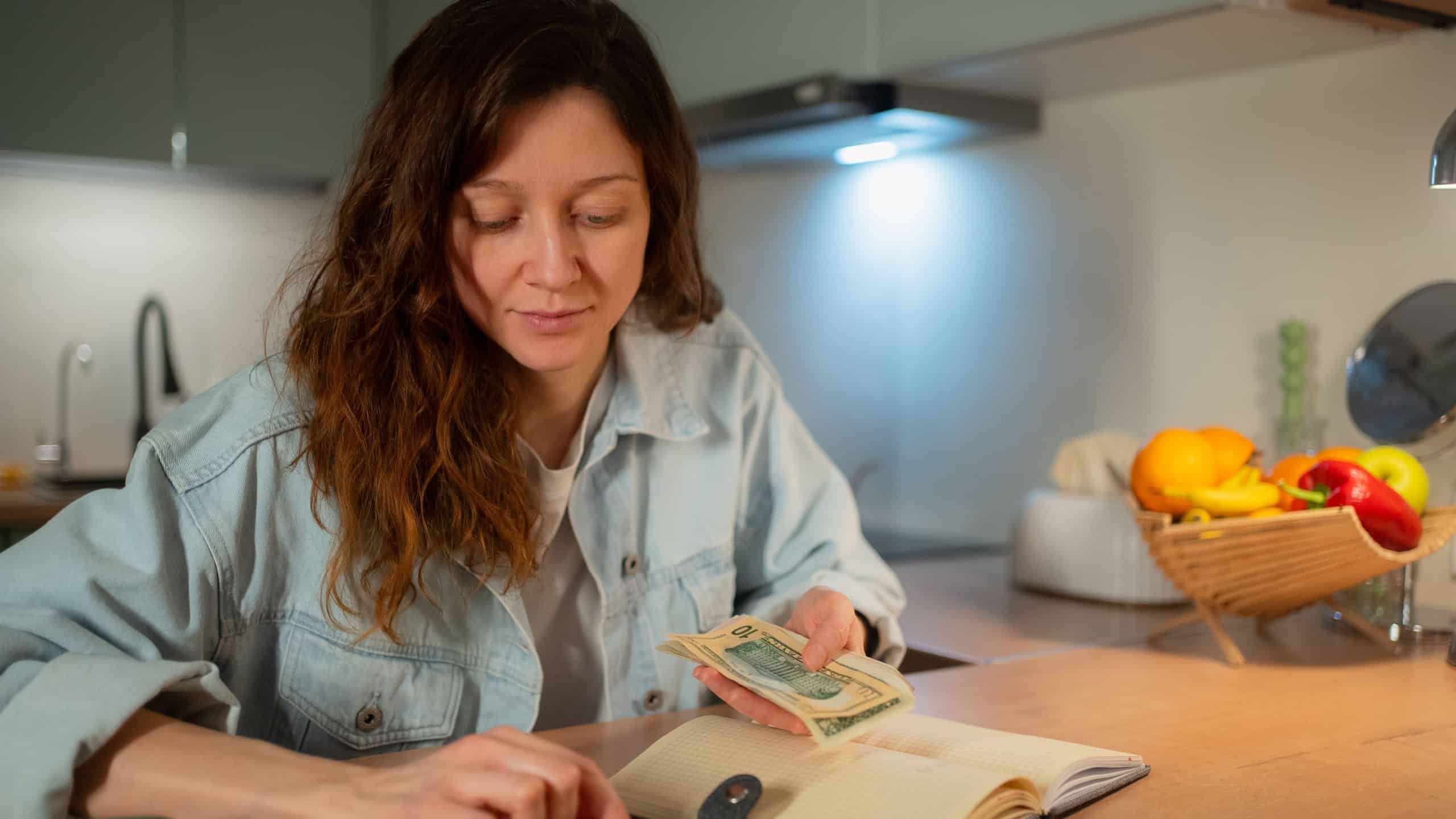 Woman calculating budget allocations and managing household expenses, holding paper money banknotes, planning utility bill payments, and organizing personal finance at home