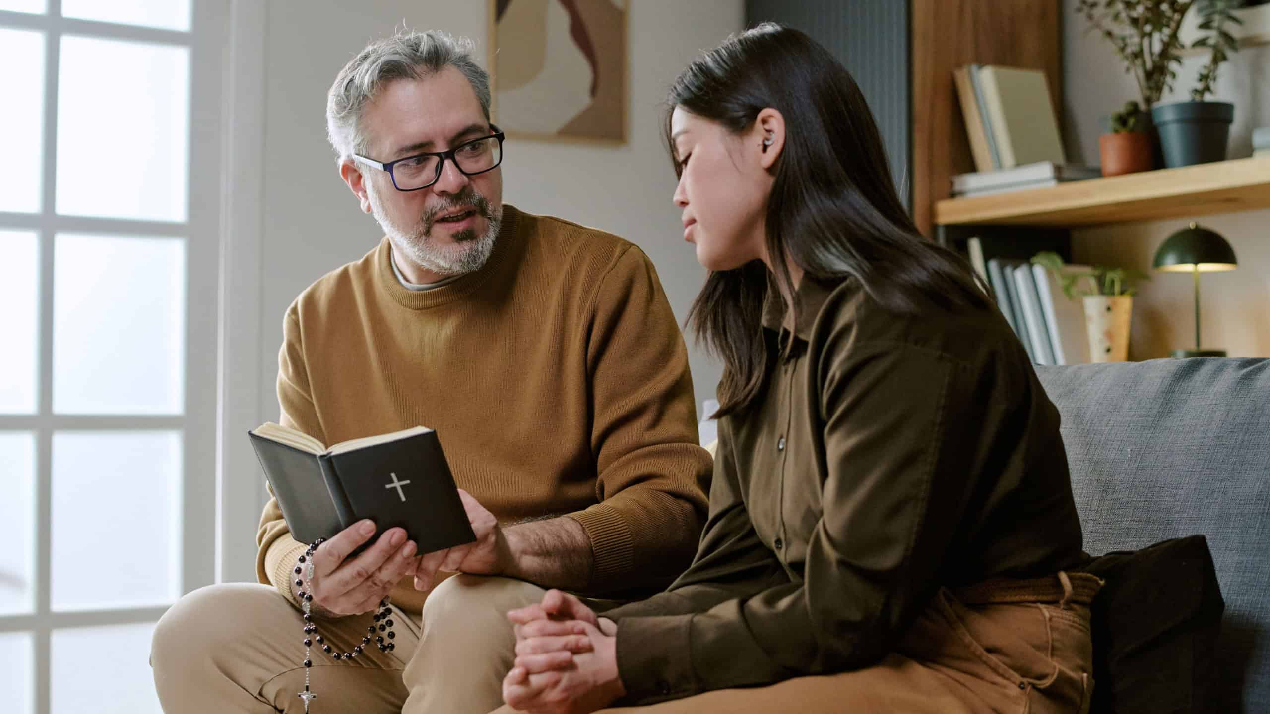 Middle aged Caucasian man holding Bible and rosary, talking to young adult woman sitting beside him on sofa, both engaged in serious conversation in home setting