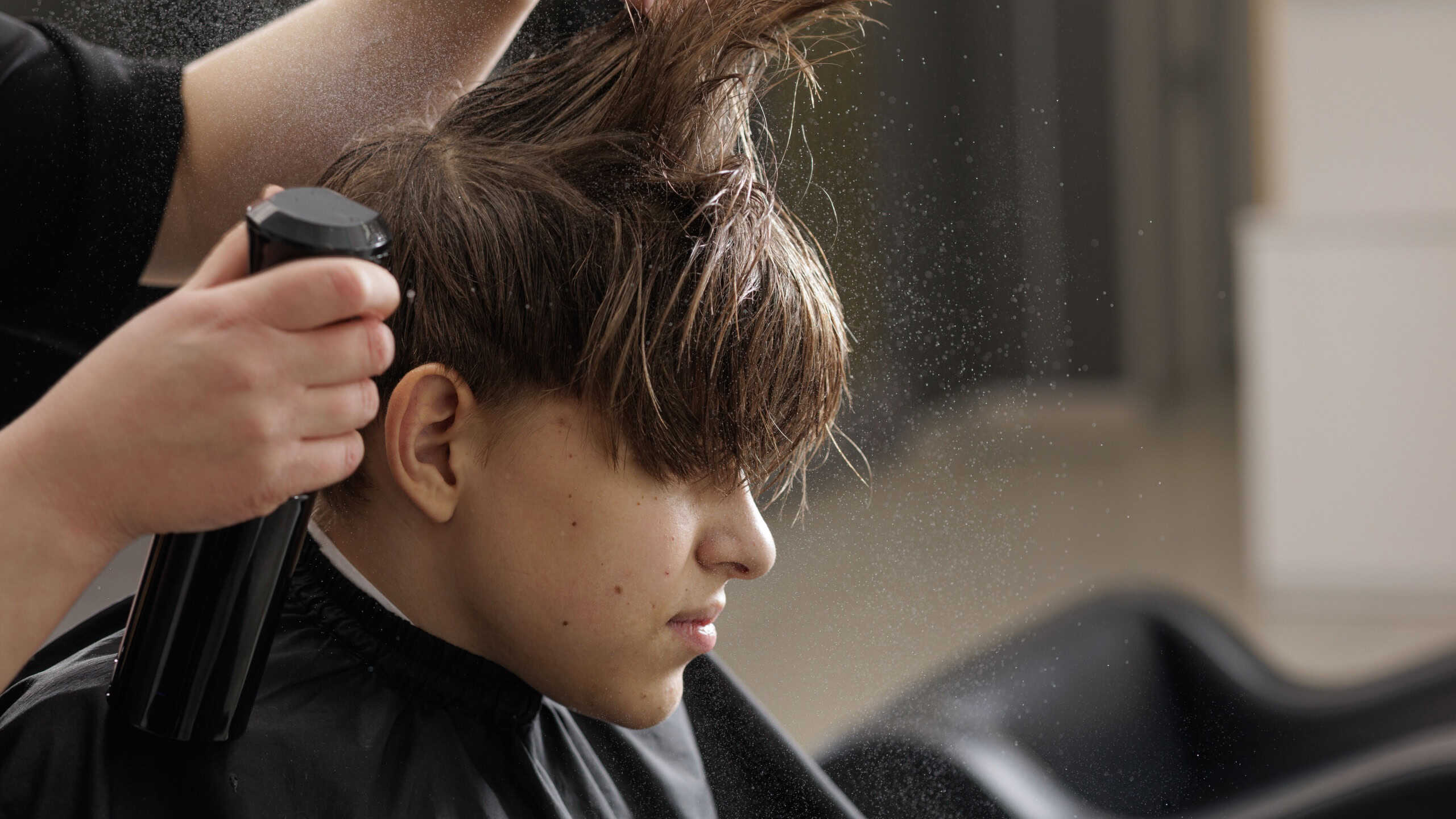 Hairdresser sprays styling product on a boy’s hair while cutting in a barbershop. Grooming, hair care routine, hygiene, and salon service concept.