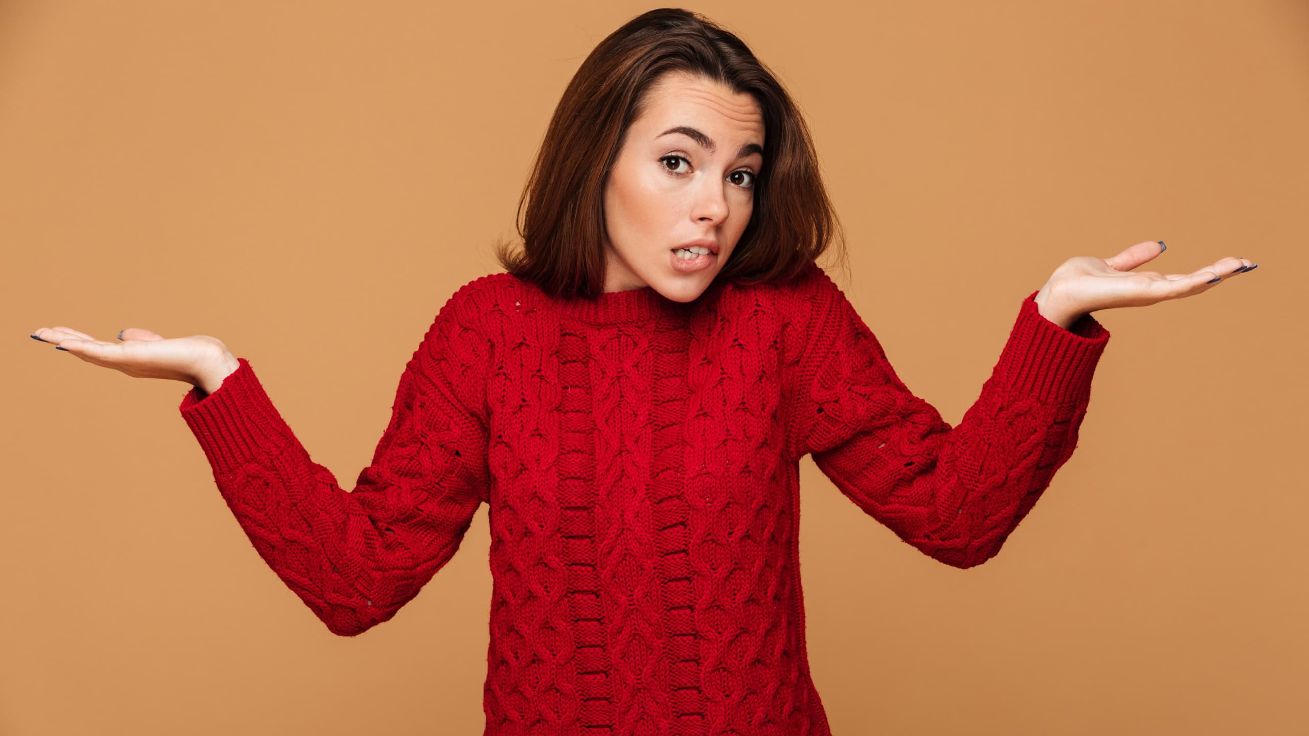 Confused young brunette woman in red knitted sweater standing with opened palms, looking at camera, isolated on beige background