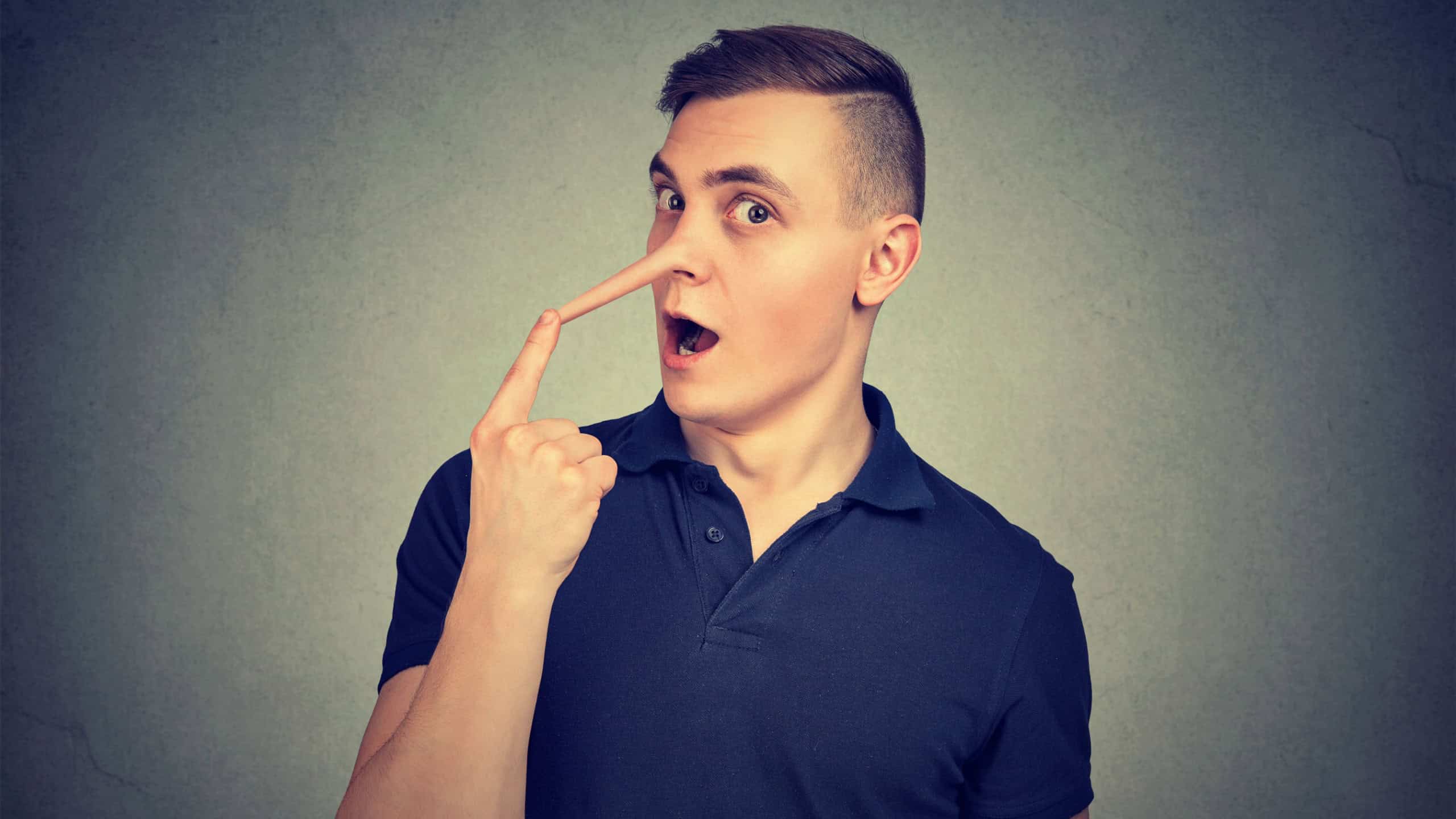 Young man looking surprised at camera touching long fake nose of liar on gray background