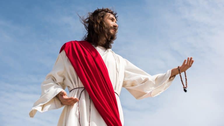 low angle view of Jesus in robe holding rosary and reaching hand against blue sky